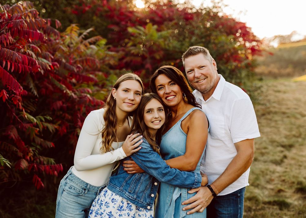 A family group poses together outdoors during golden hour with red autumn foliage behind them.