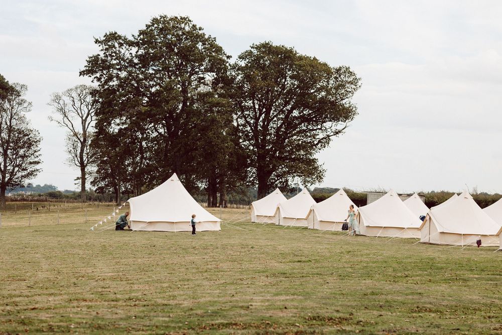 Row of white canvas bell tents set up in a grassy field with tall trees in the background.