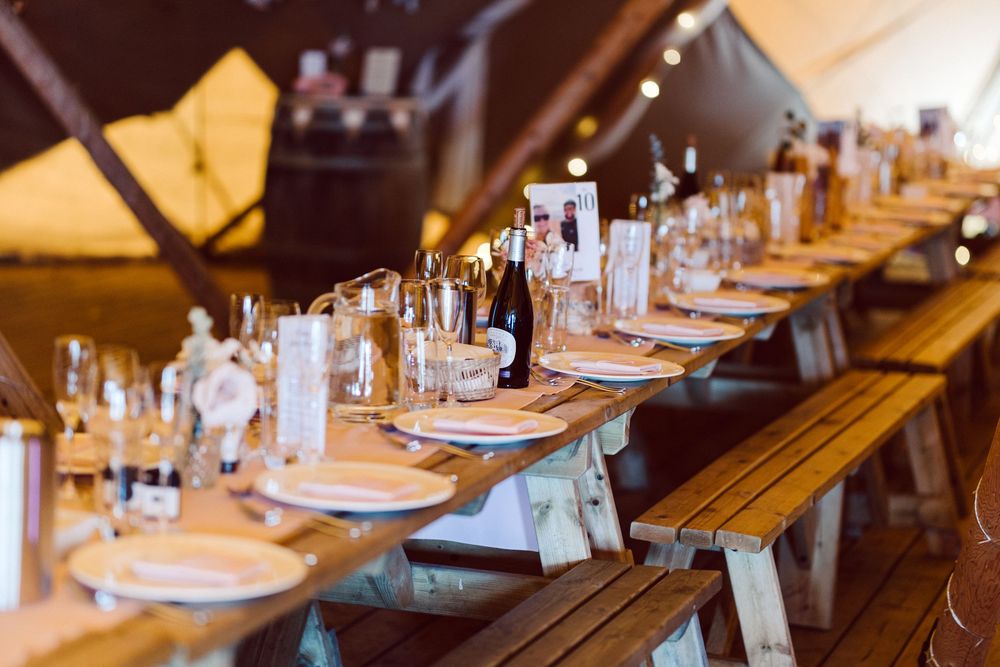 Long wooden table set for a rustic outdoor wedding reception with elegant place settings and glassware.