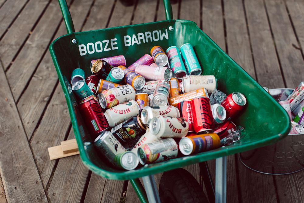 Green wheelbarrow filled with empty beverage cans and bottles on wooden deck.