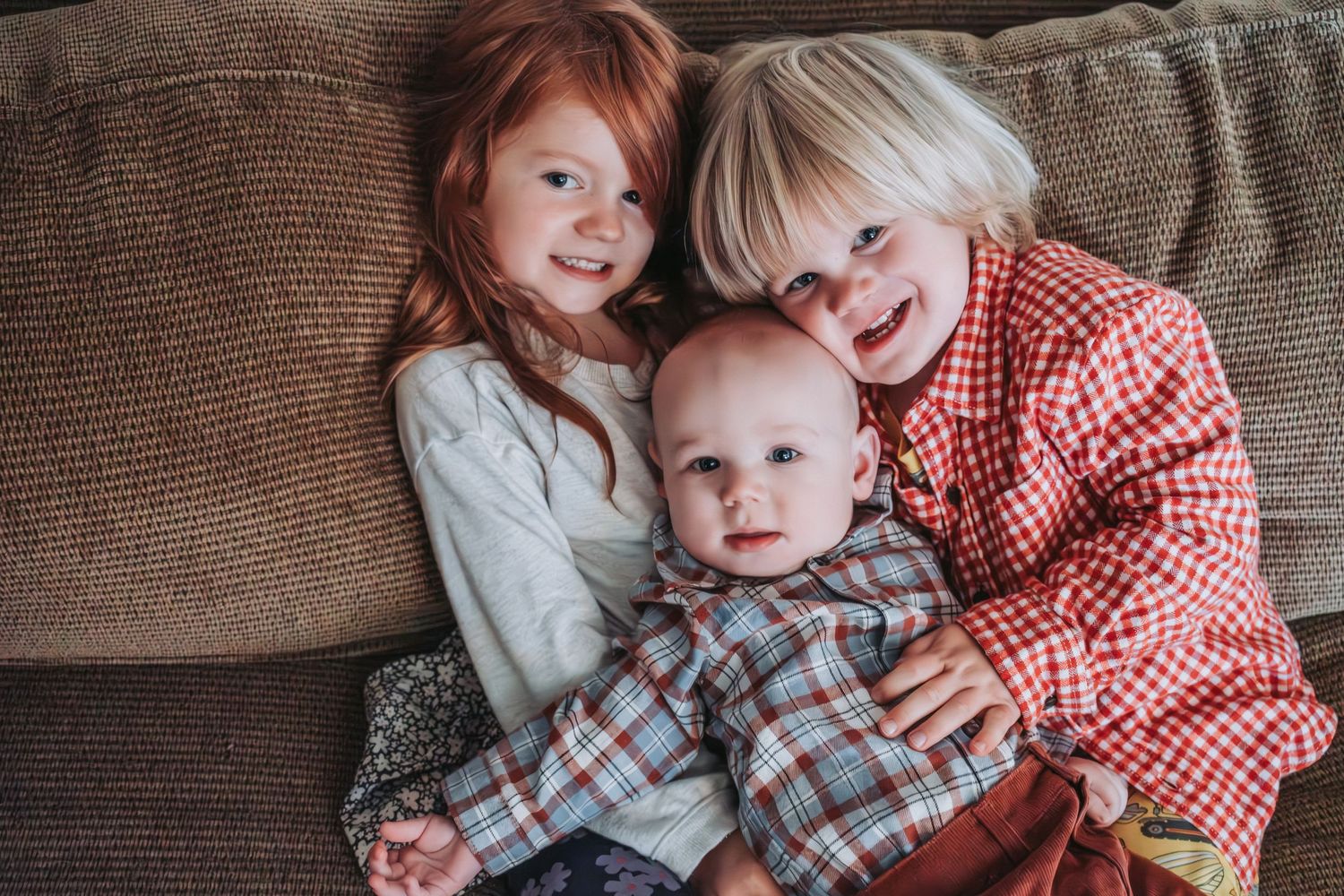 Children in coordinating plaid and gingham outfits cuddle and laugh together on a brown couch.