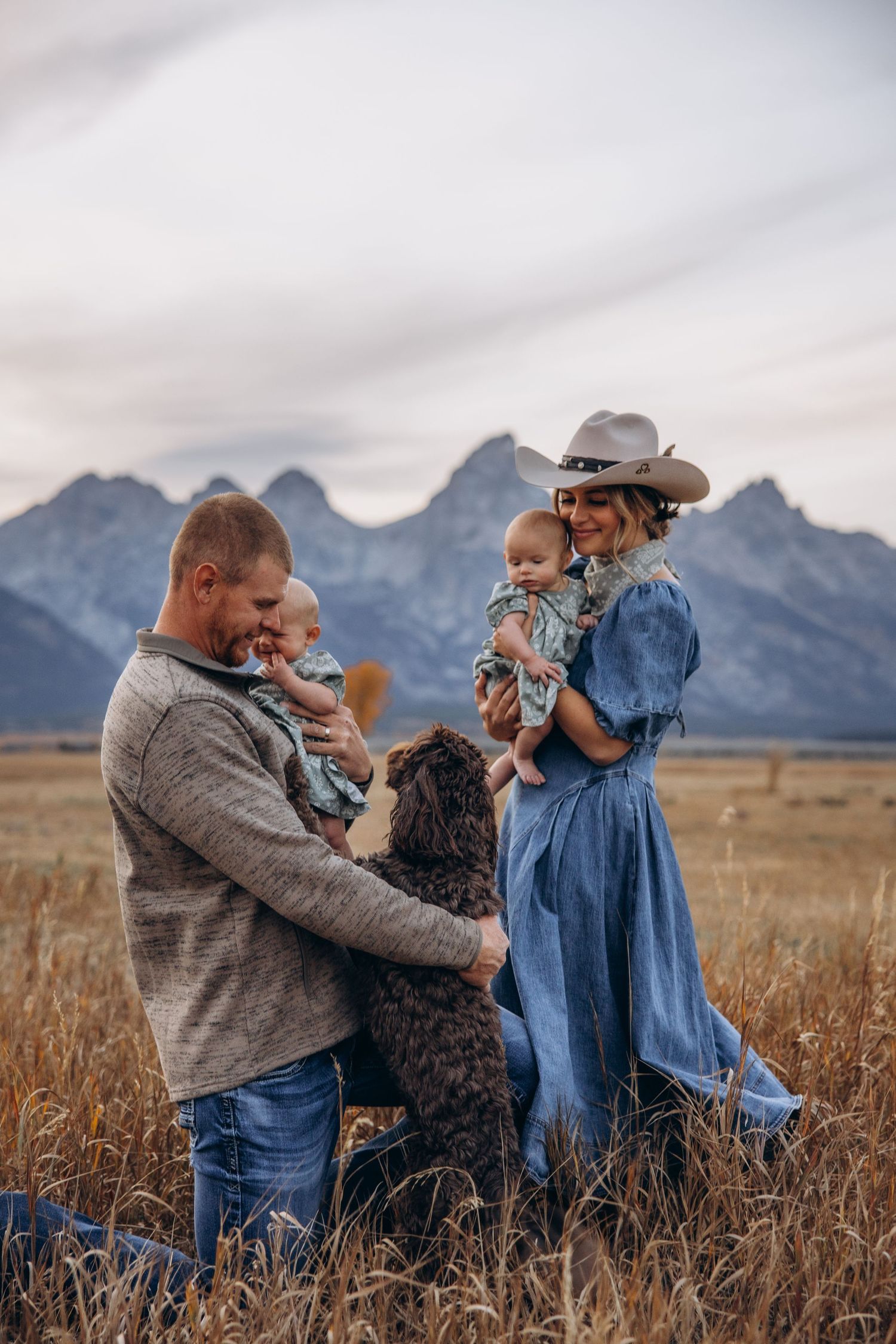 Family and their curly-haired dog enjoy a sunset moment in a field with majestic mountain peaks in the background.