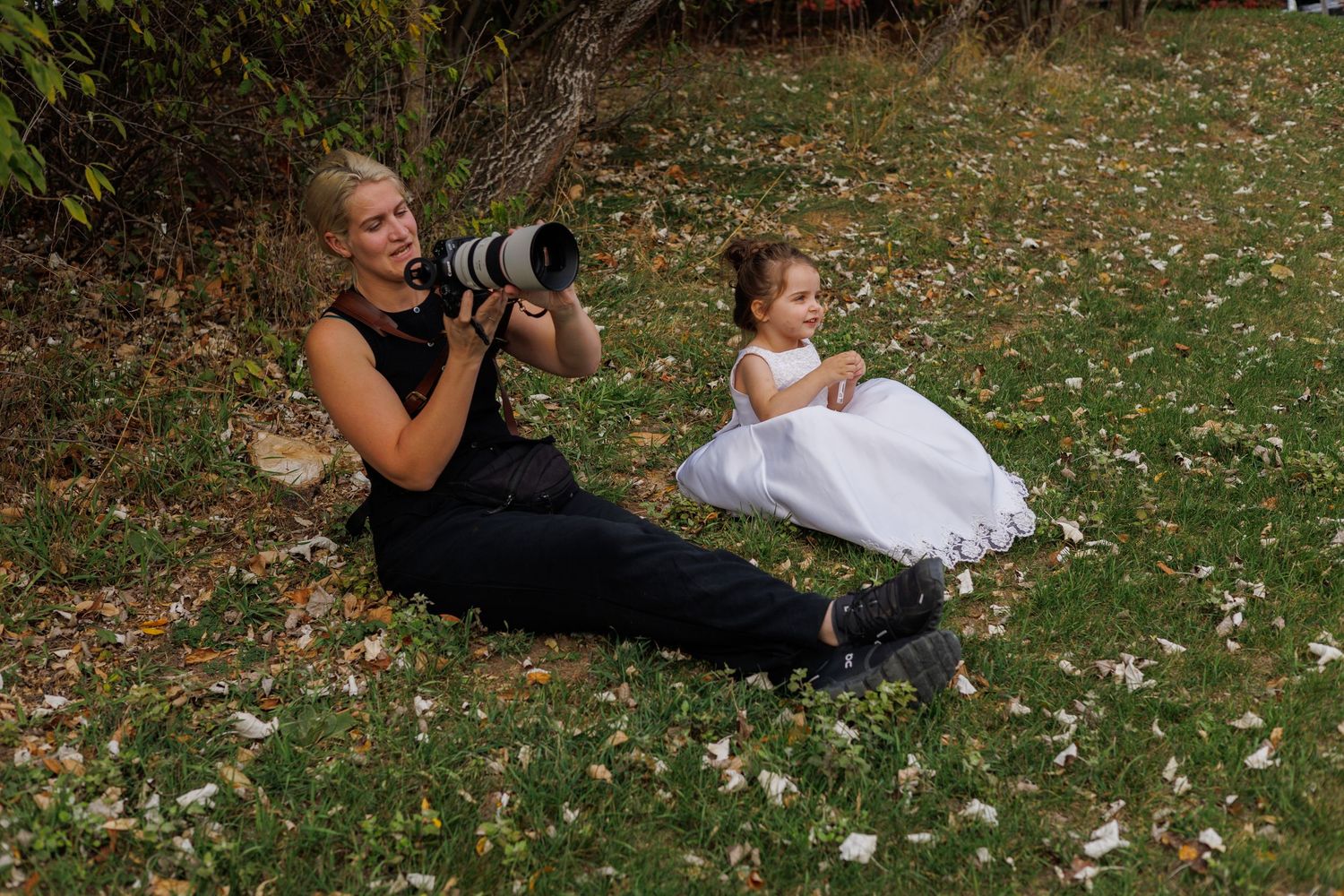 A photographer lies on grass taking pictures while a small child in a flower girl sits nearby during a wedding shoot.