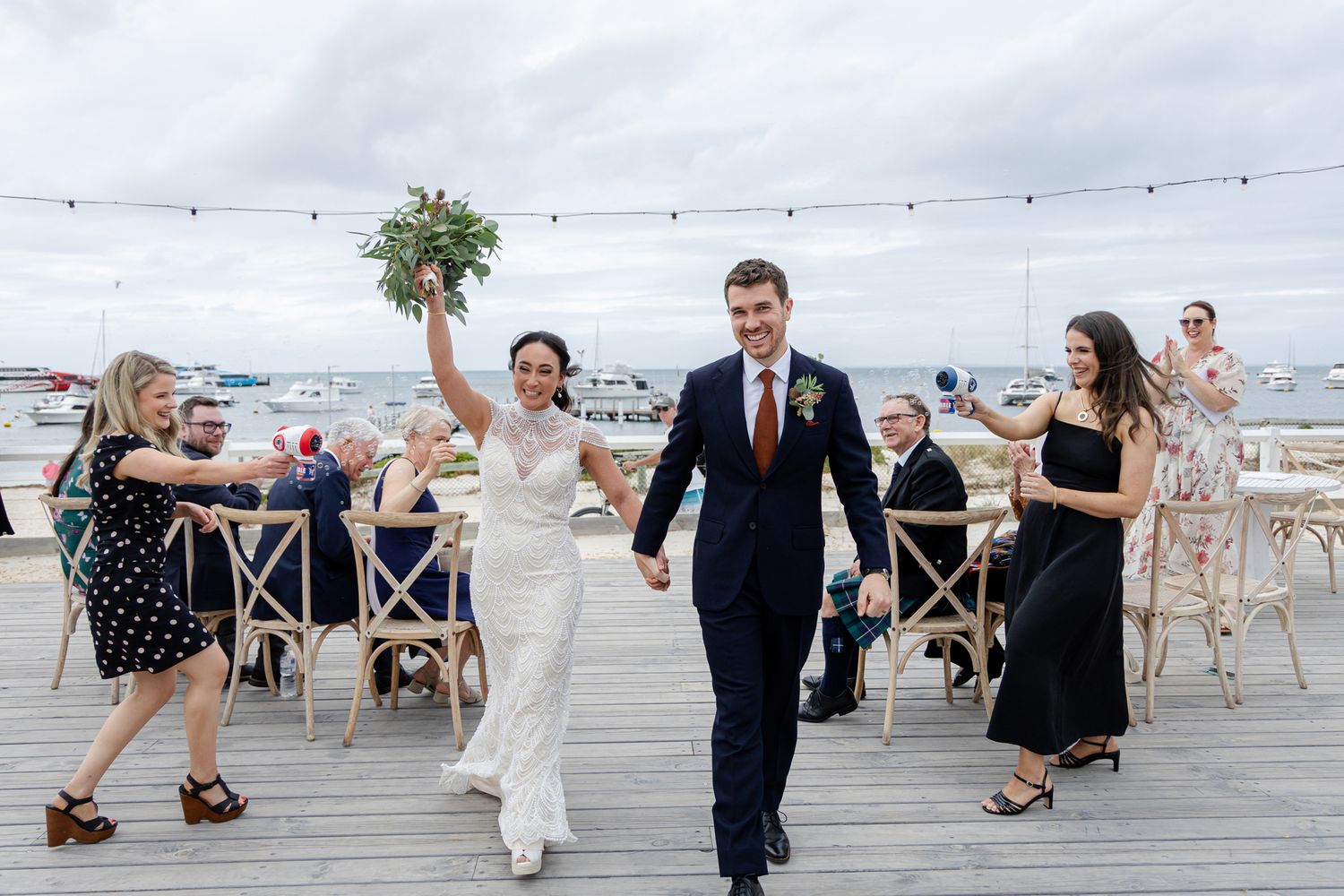 Wedding guests dance together on a wooden deck overlooking the beach during a sunset celebration.