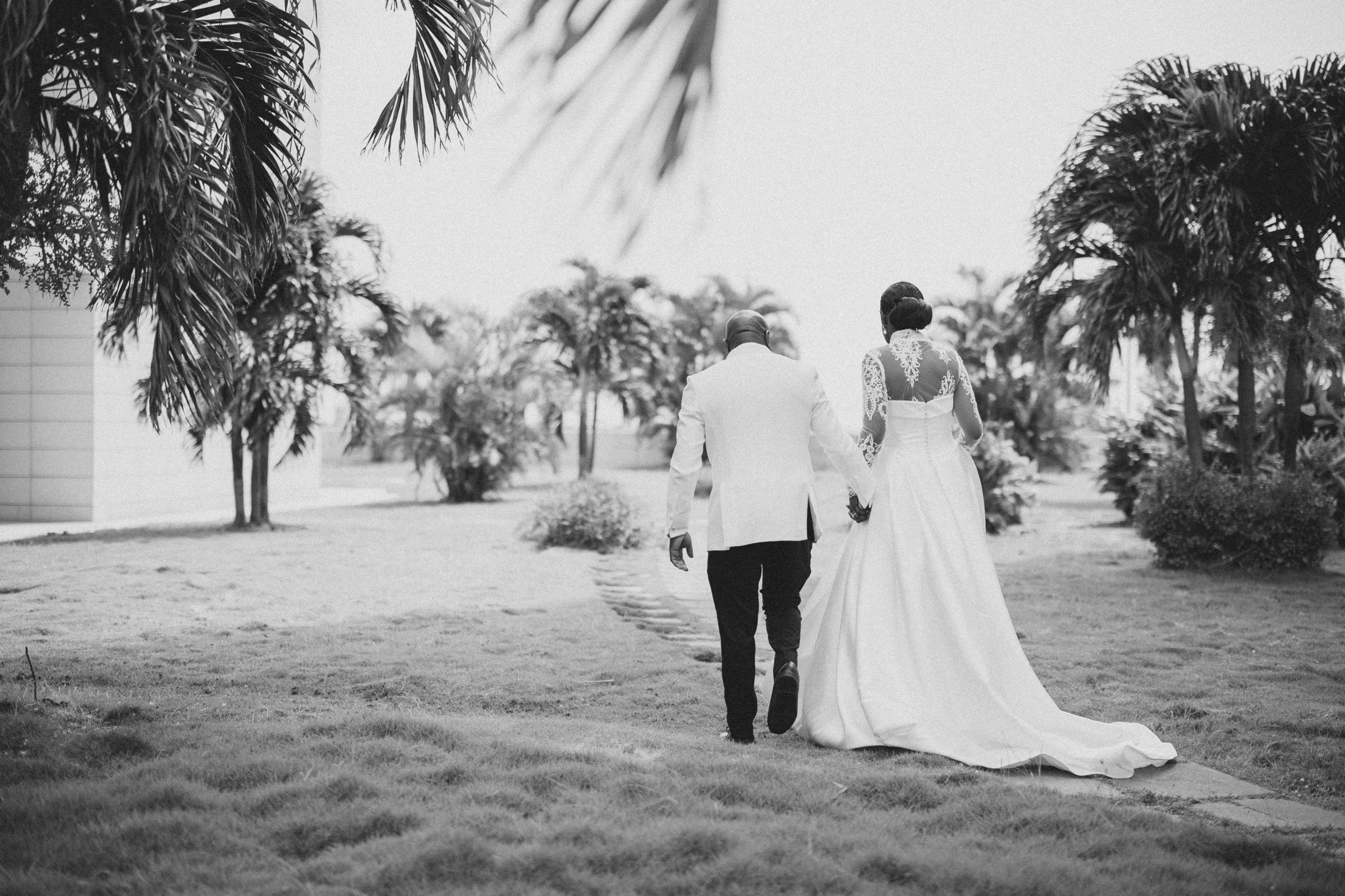 A couple in wedding attire walks through a palm tree lined path in black and white photography.