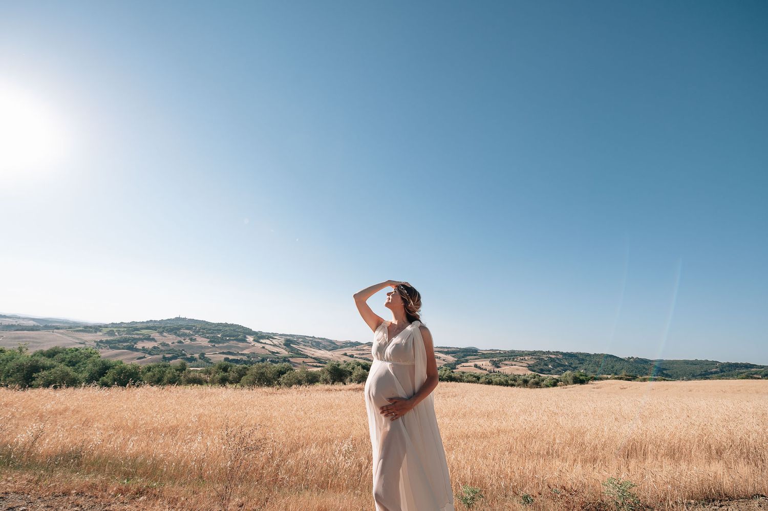 Woman in flowing white dress stands in golden field during sunset with mountains in background.
