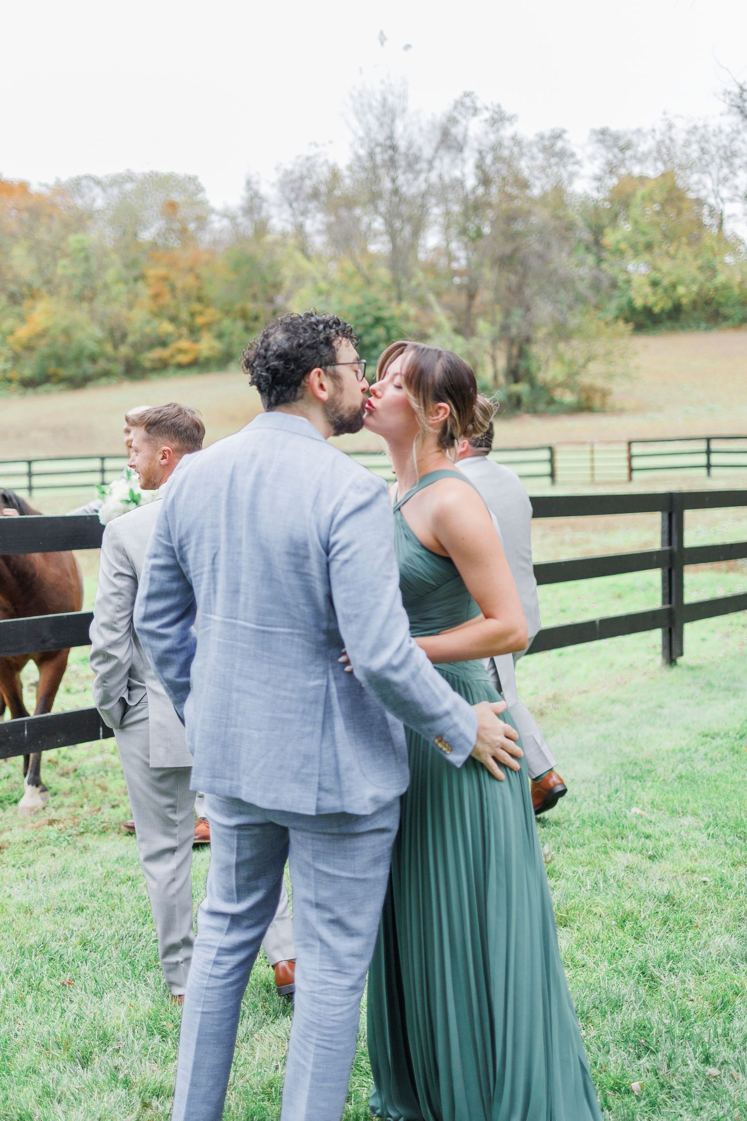 A couple in wedding attire share an intimate moment in a grassy field with wooden fencing and autumn trees in background.