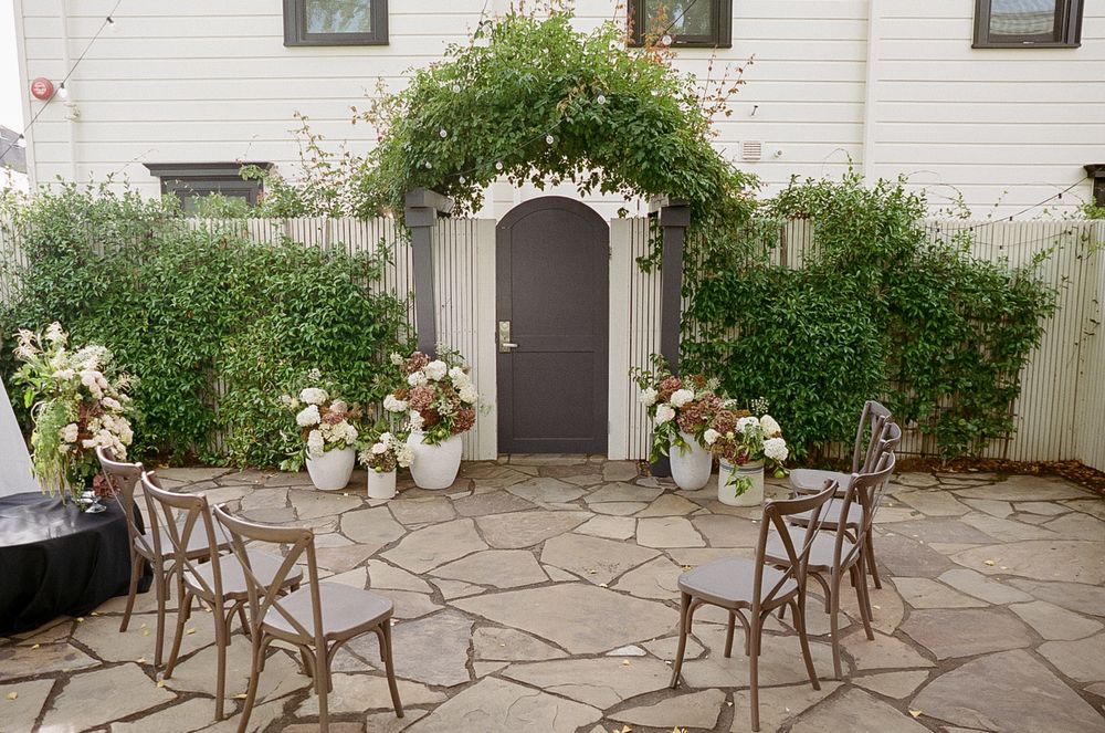 Stone patio courtyard with wooden chairs and floral arrangements against a white building exterior.