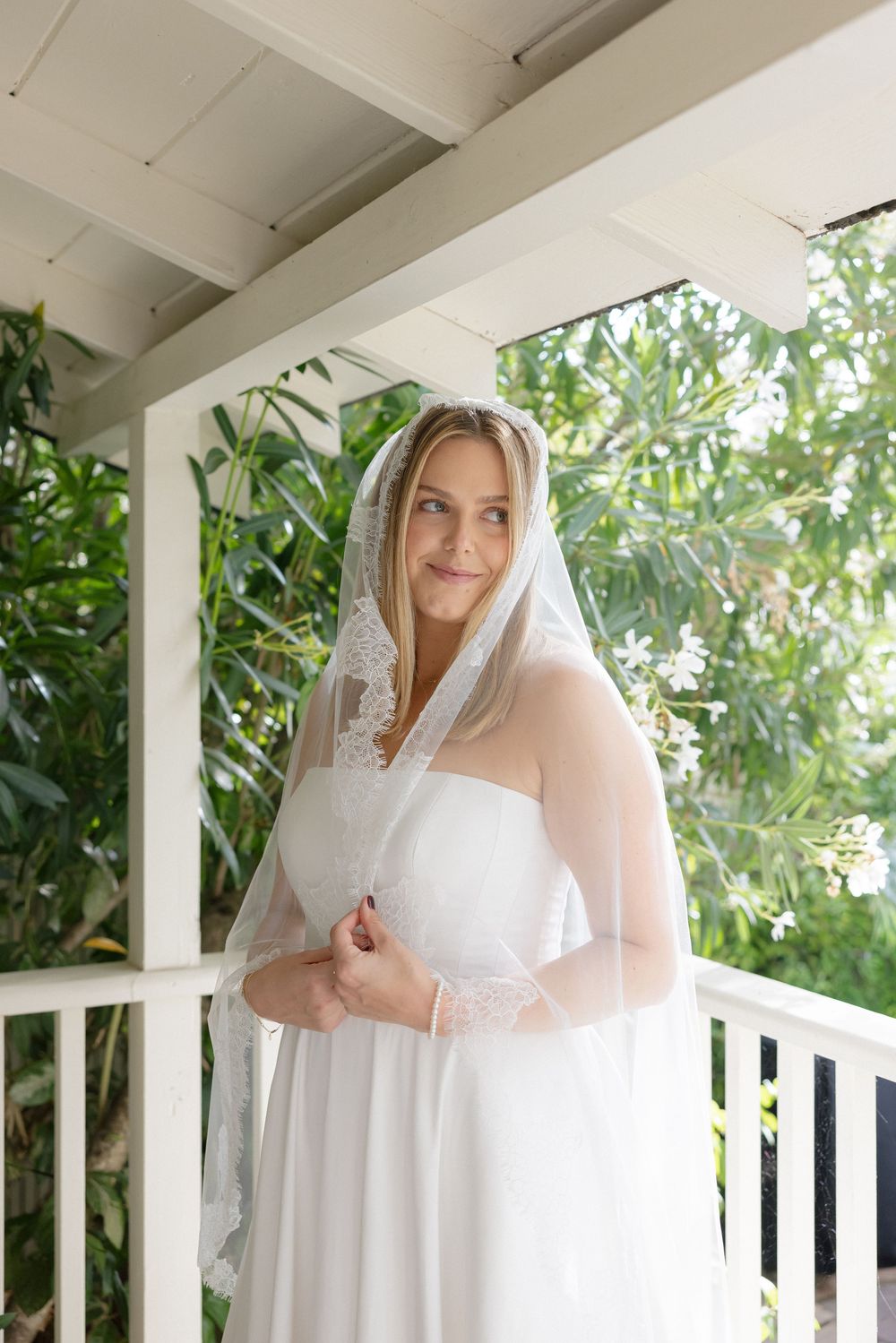 A bride in a classic white wedding dress poses on a porch surrounded by greenery.