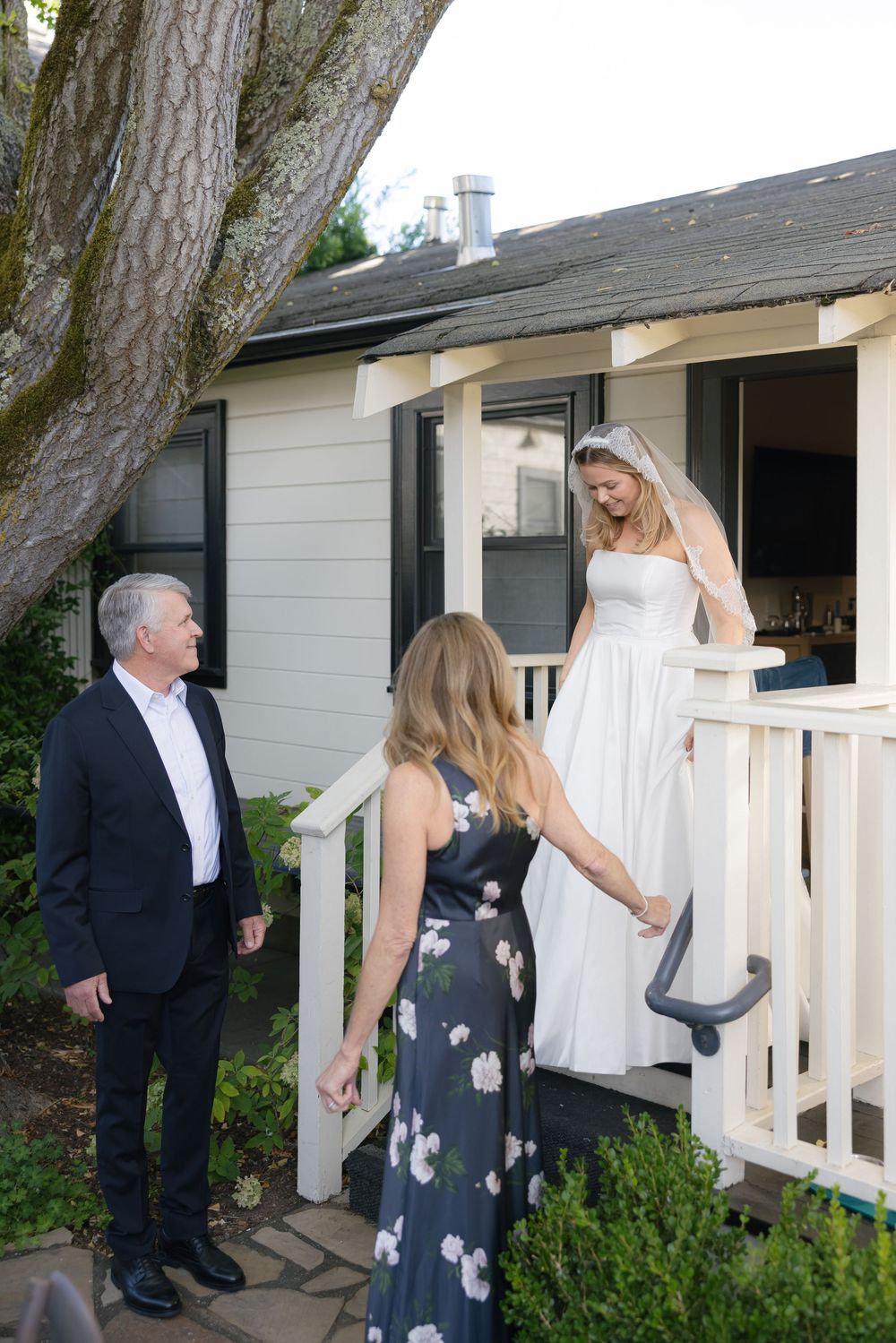 Wedding party interacts on white porch with lush garden surroundings.