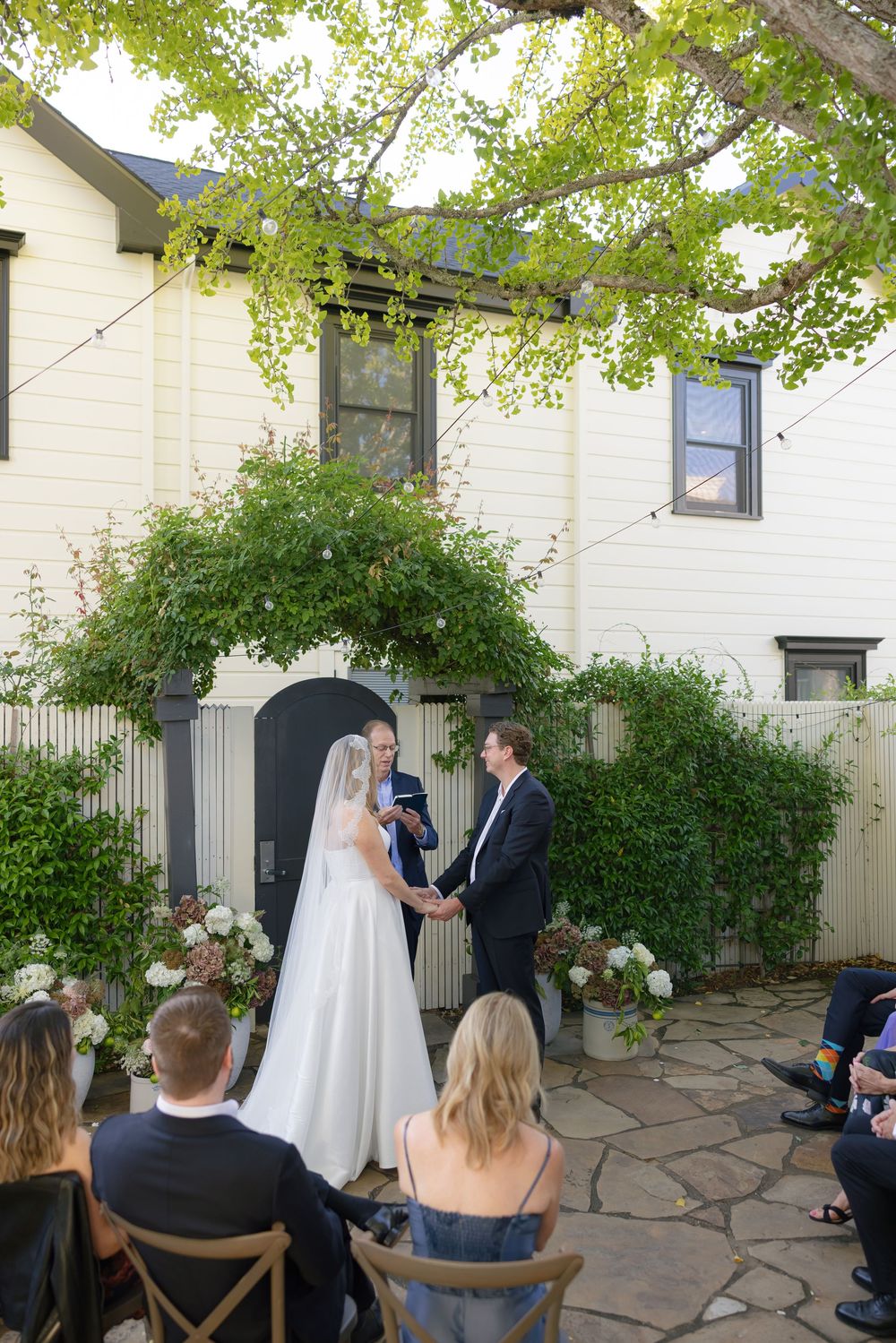 Outdoor wedding ceremony with guests seated on wooden chairs under green leafy trees.