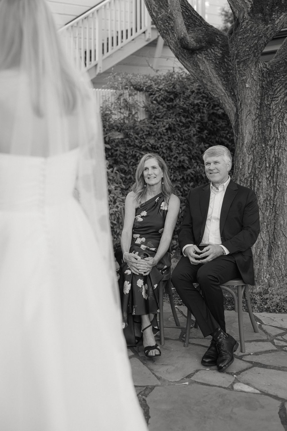 A couple sits together on a bench at a formal outdoor event in black and white photography.