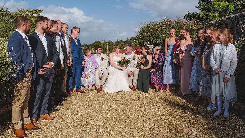 Wedding guests gather outdoors on a sunny day for a panoramic group photo against a scenic backdrop.