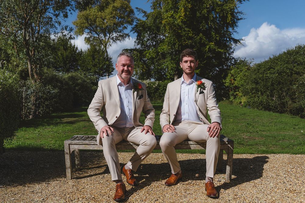 A group in light-colored suits and brown shoes pose together on a gravel path in a scenic outdoor setting.