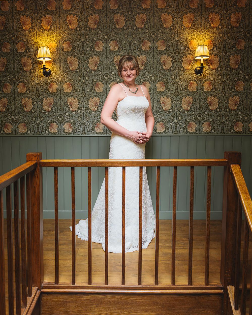 A person in a white wedding dress stands elegantly on a staircase landing with vintage wallpaper and sconces in the background.