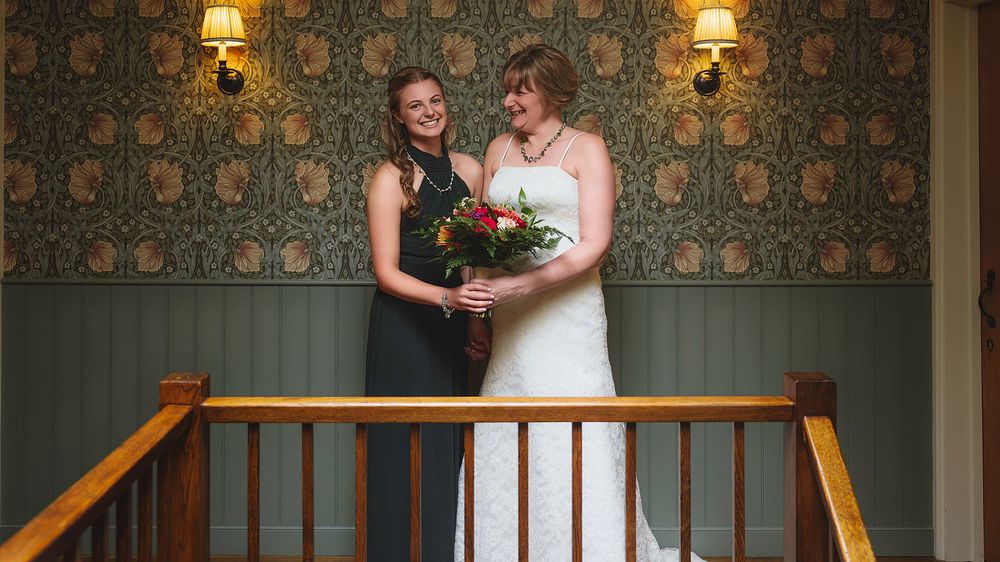 Bride in white wedding dress stands on ornate staircase with green floral wallpaper and warm lighting.