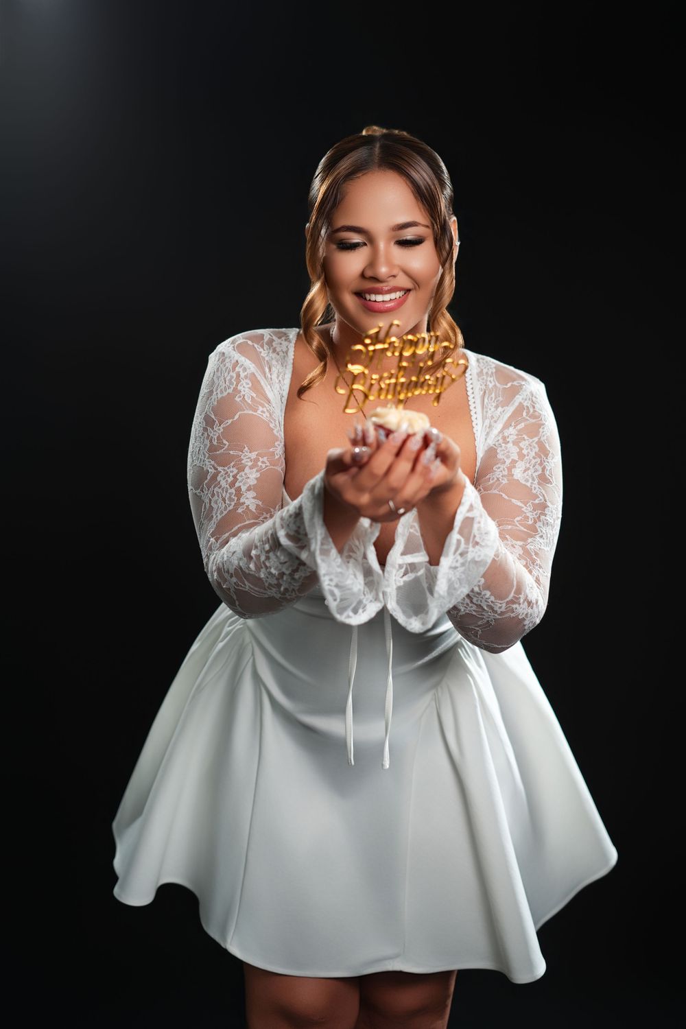 Woman in white lace dress poses playfully with birthday cake topper against black background.