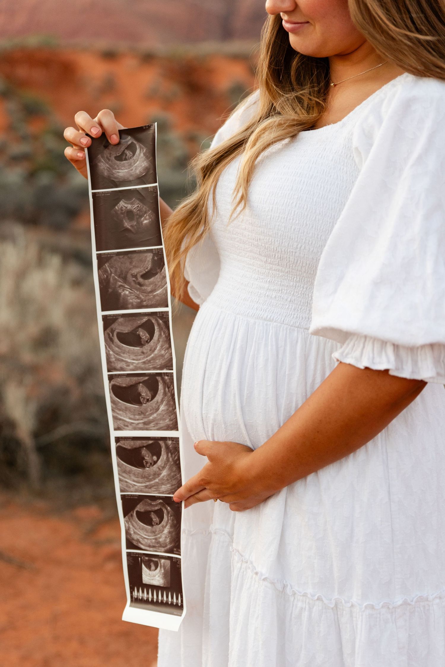 closeup of a pregnant moms belly in a white dress holding a sonogram