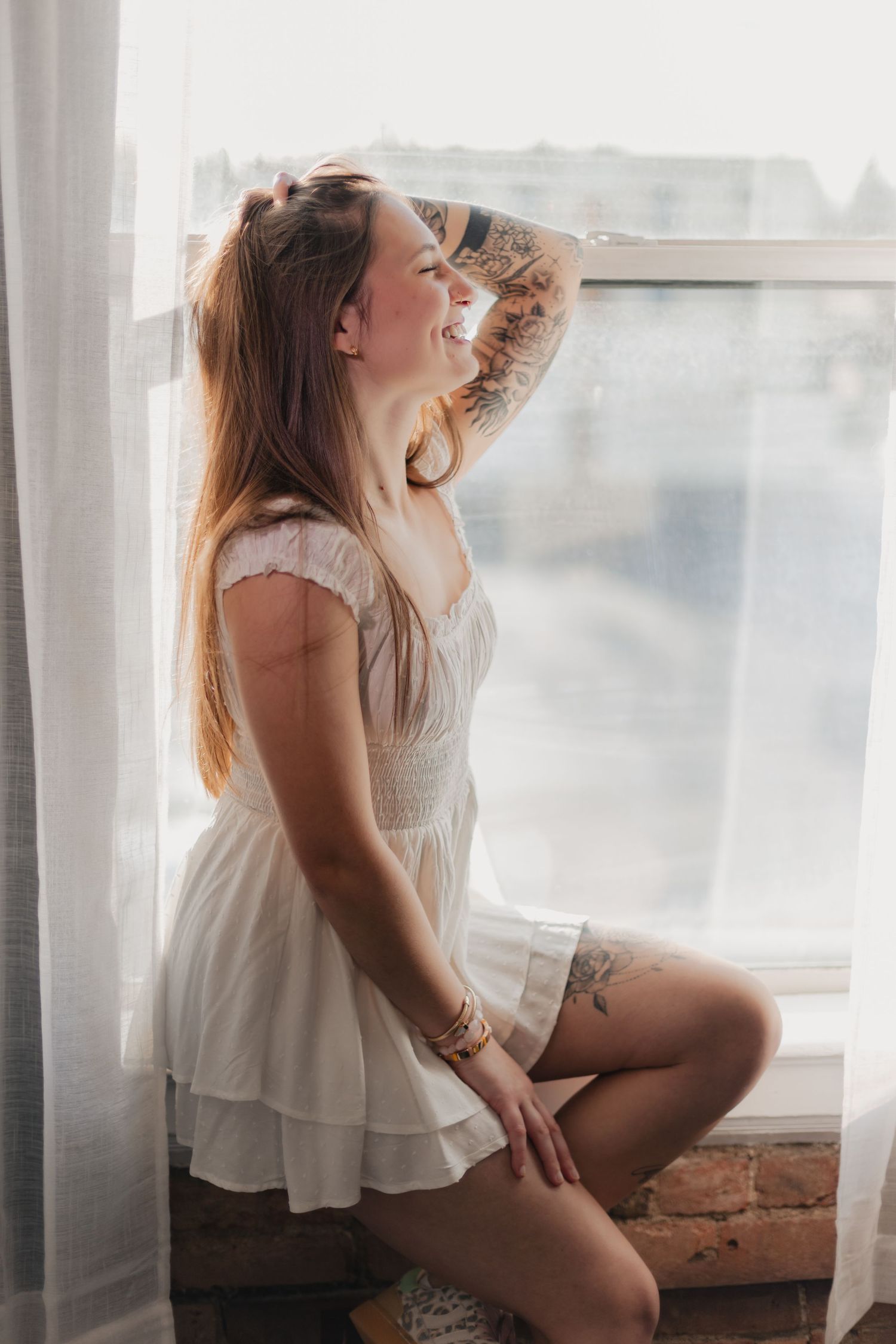 Woman in white top poses artistically near window in soft natural light creating dreamy atmosphere.