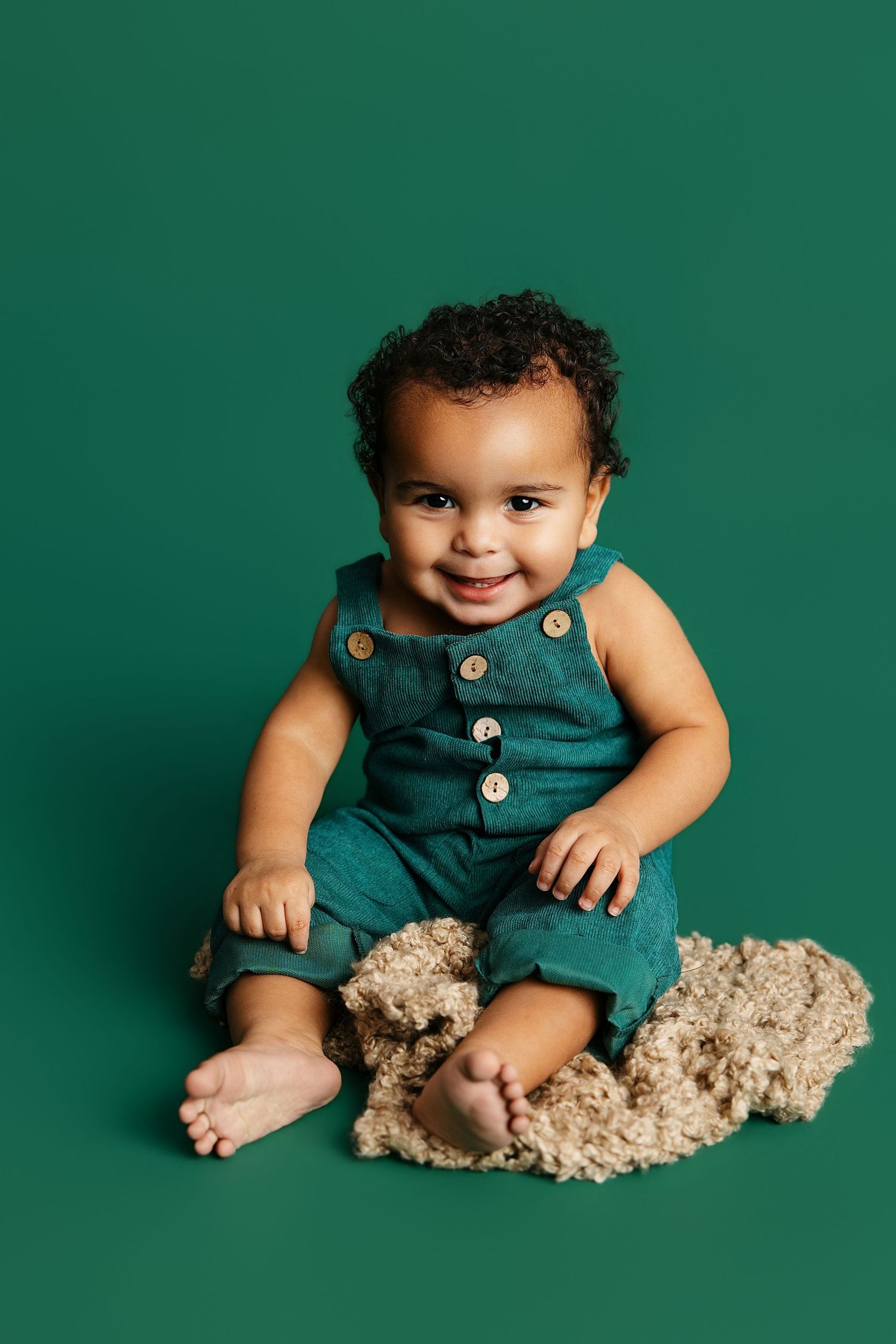 Baby wearing green overalls sits against emerald backdrop with plush teddy bear in studio setting.