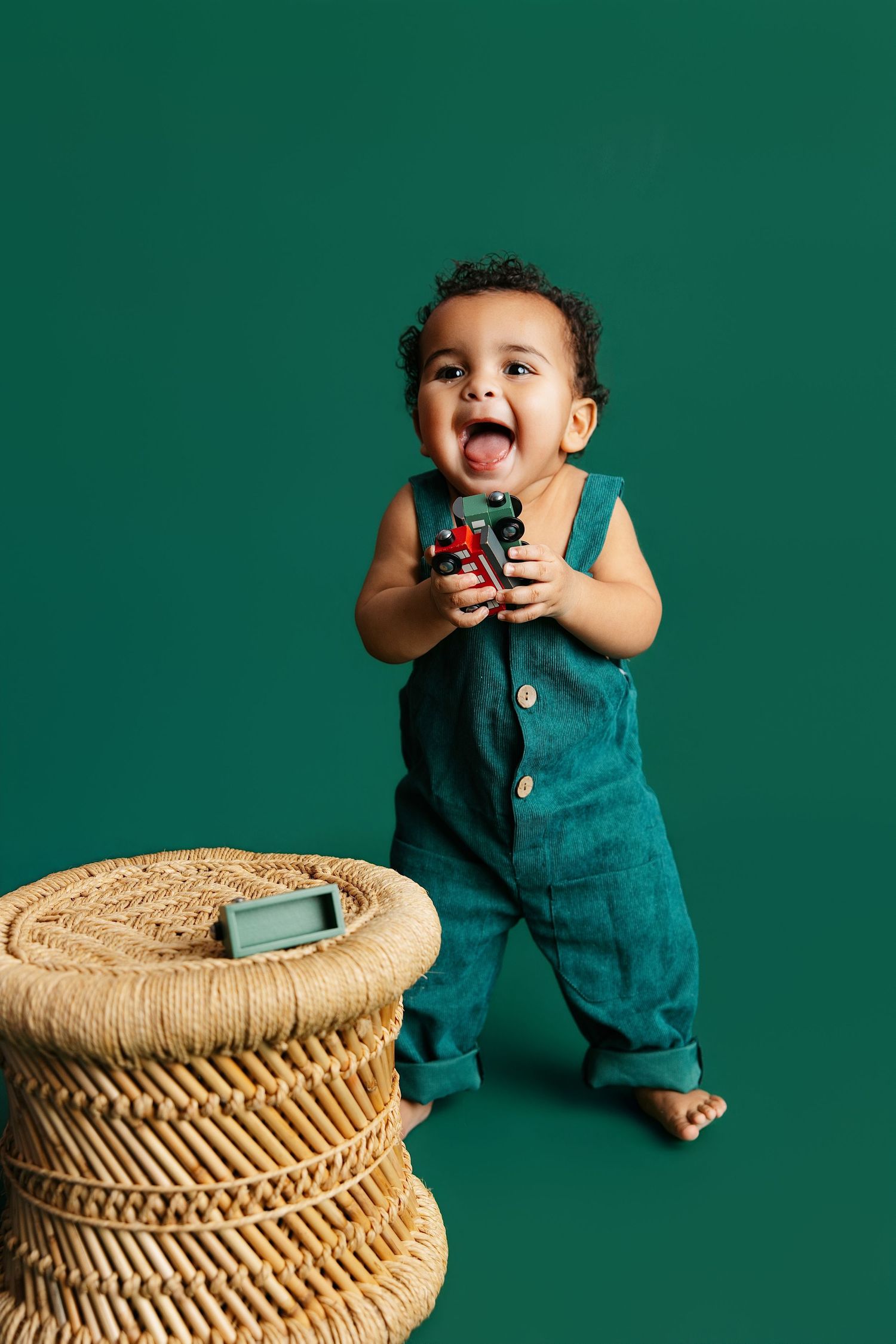 A joyful toddler in teal overalls plays near a woven basket against an emerald green backdrop.