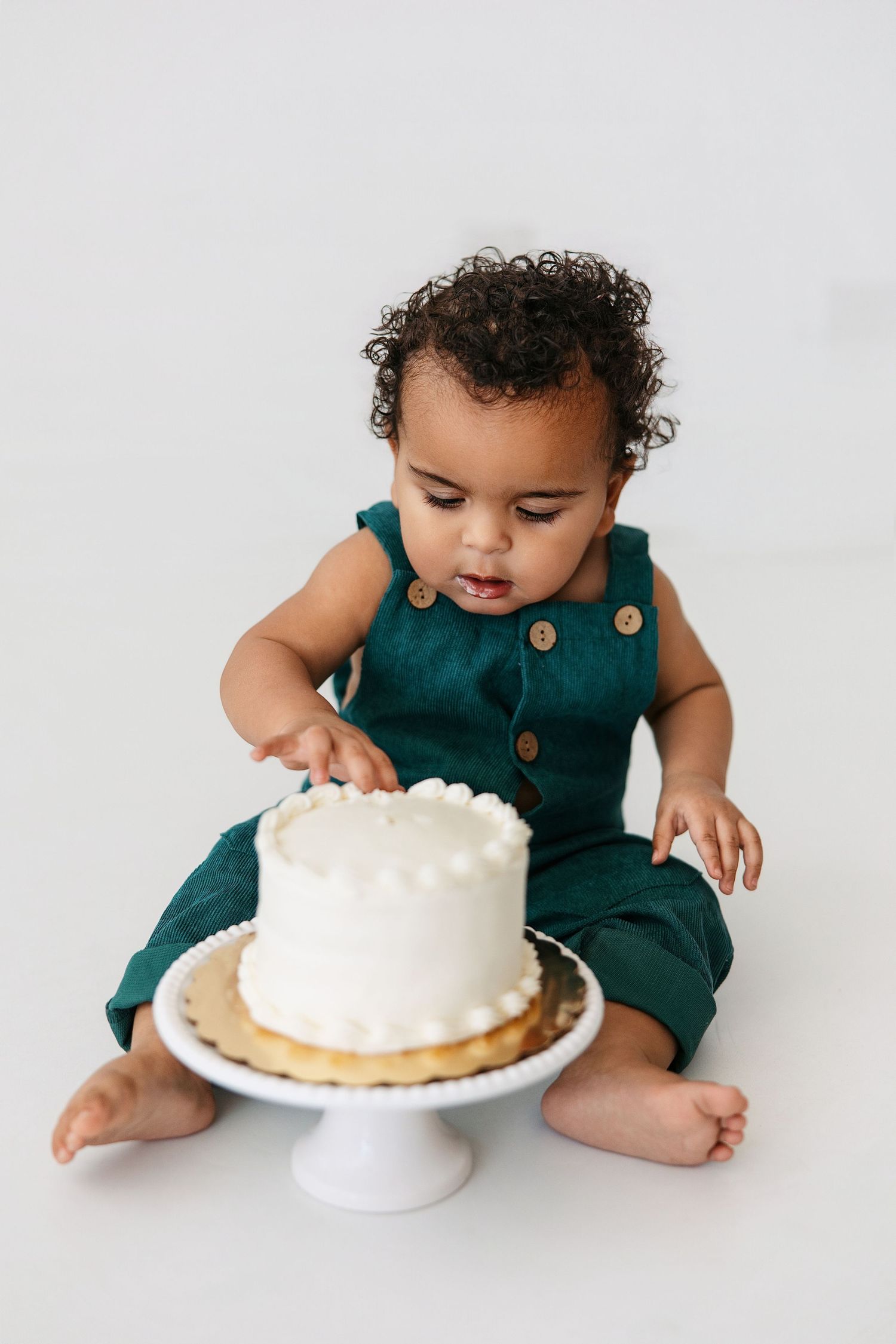 Small child in teal romper reaching for white frosted cake on pedestal against white background.
