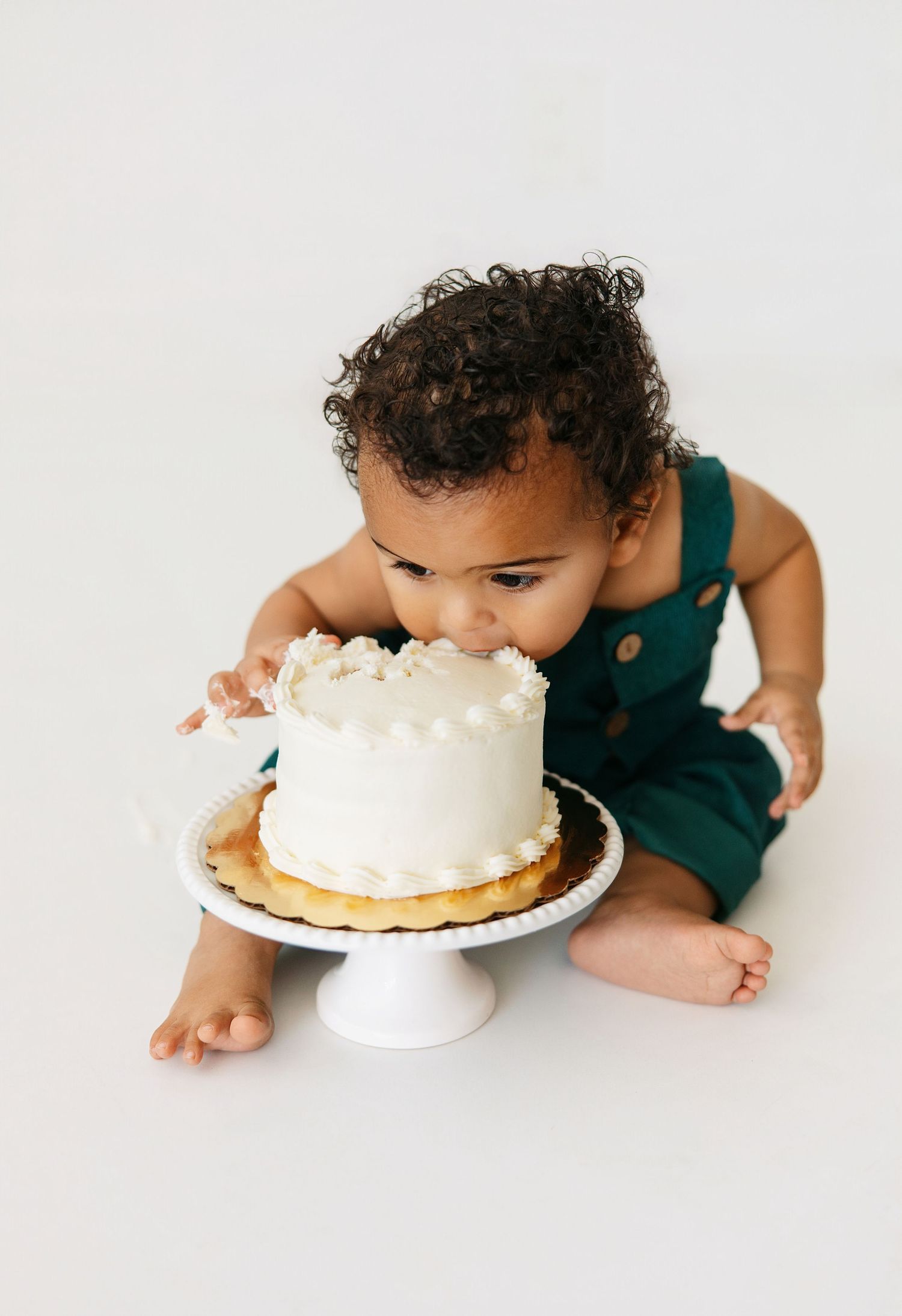 A sequence of photos showing a small child in teal clothing taking bites of a white frosted cake on a cake stand.