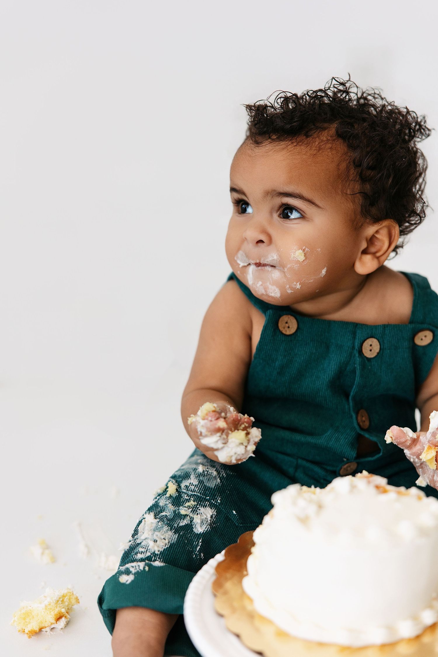 A messy cake smash photo session shows a toddler enjoying their birthday treat against a white background.