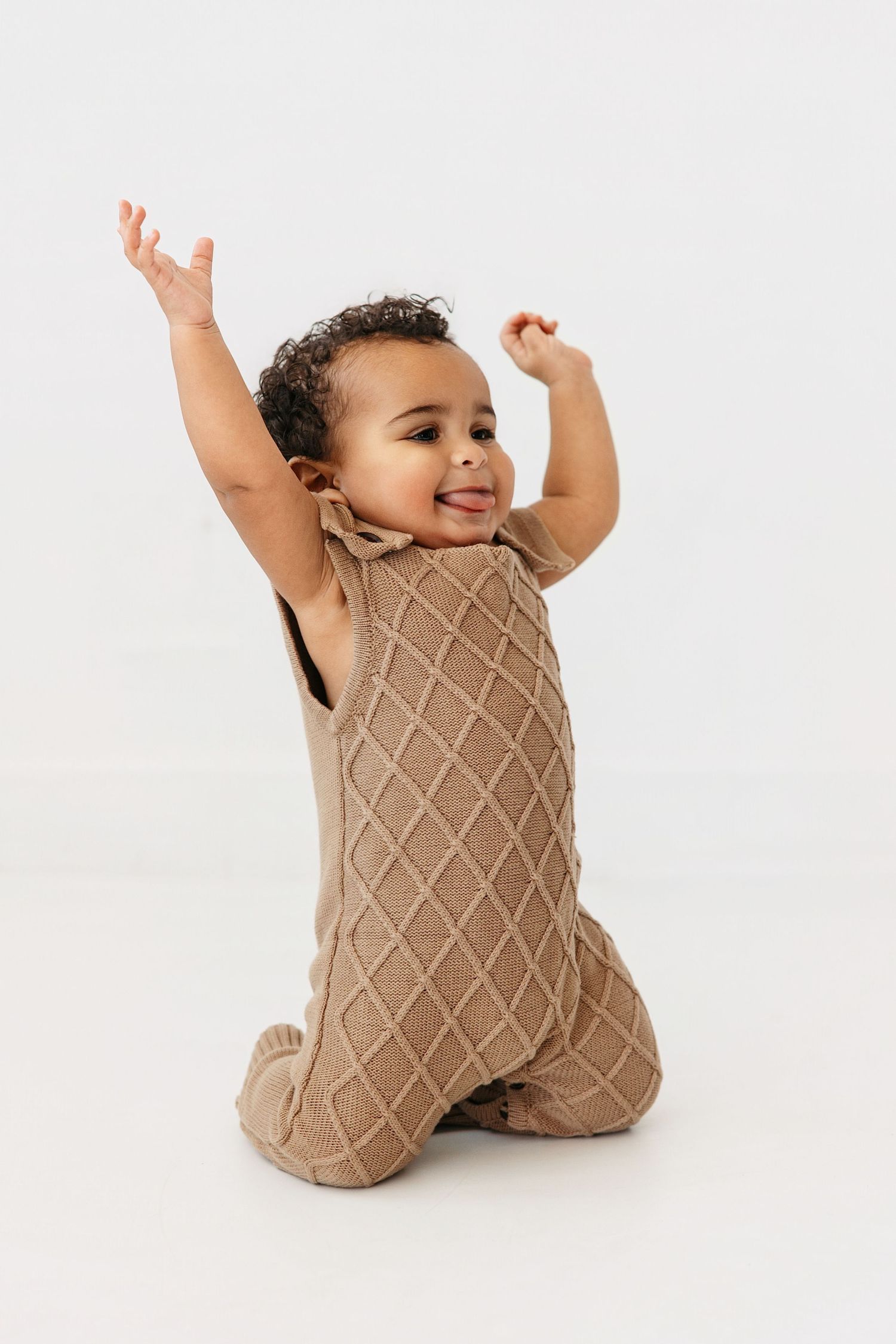 Baby in tan knit romper sitting and raising arms joyfully against white background.
