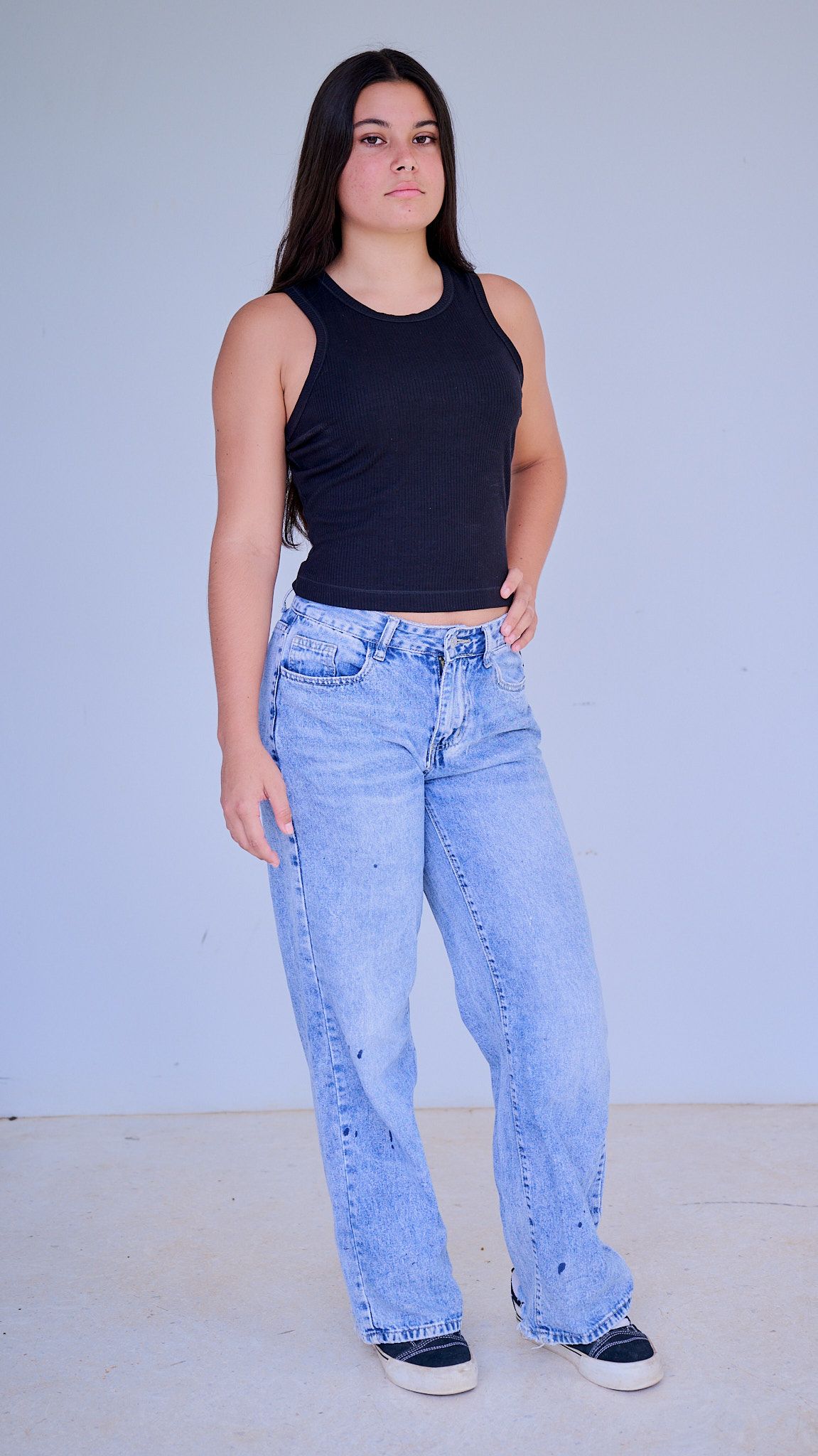 Model wearing a white crop top and light blue high-waisted jeans poses against a light blue background.