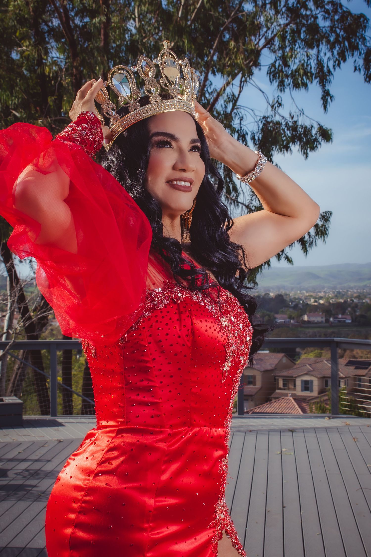A figure in a flowing red lace dress poses gracefully on a wooden deck with trees and cityscape in the background.