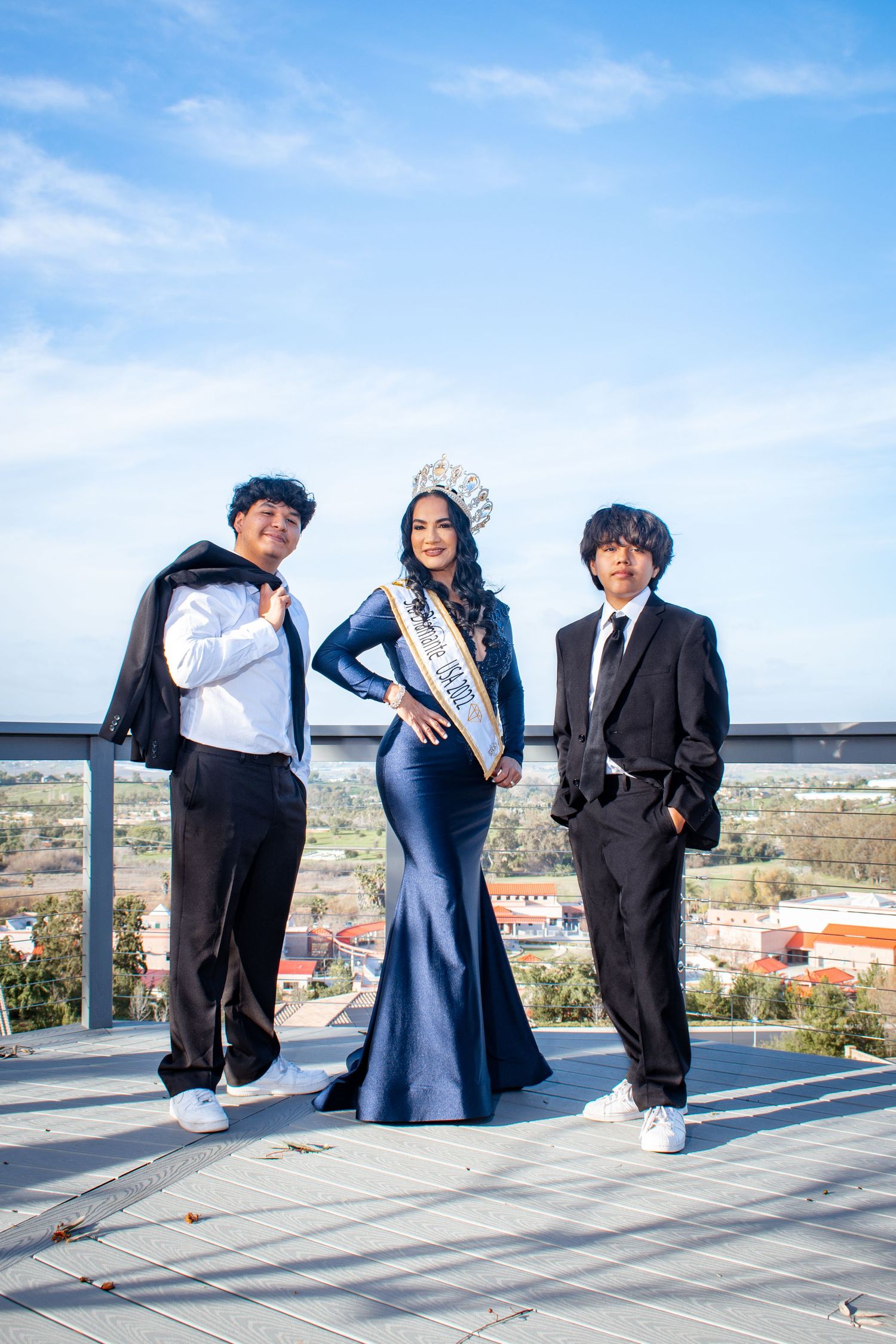 Three people in formal attire pose playfully on a rooftop terrace with a city skyline view in the background.