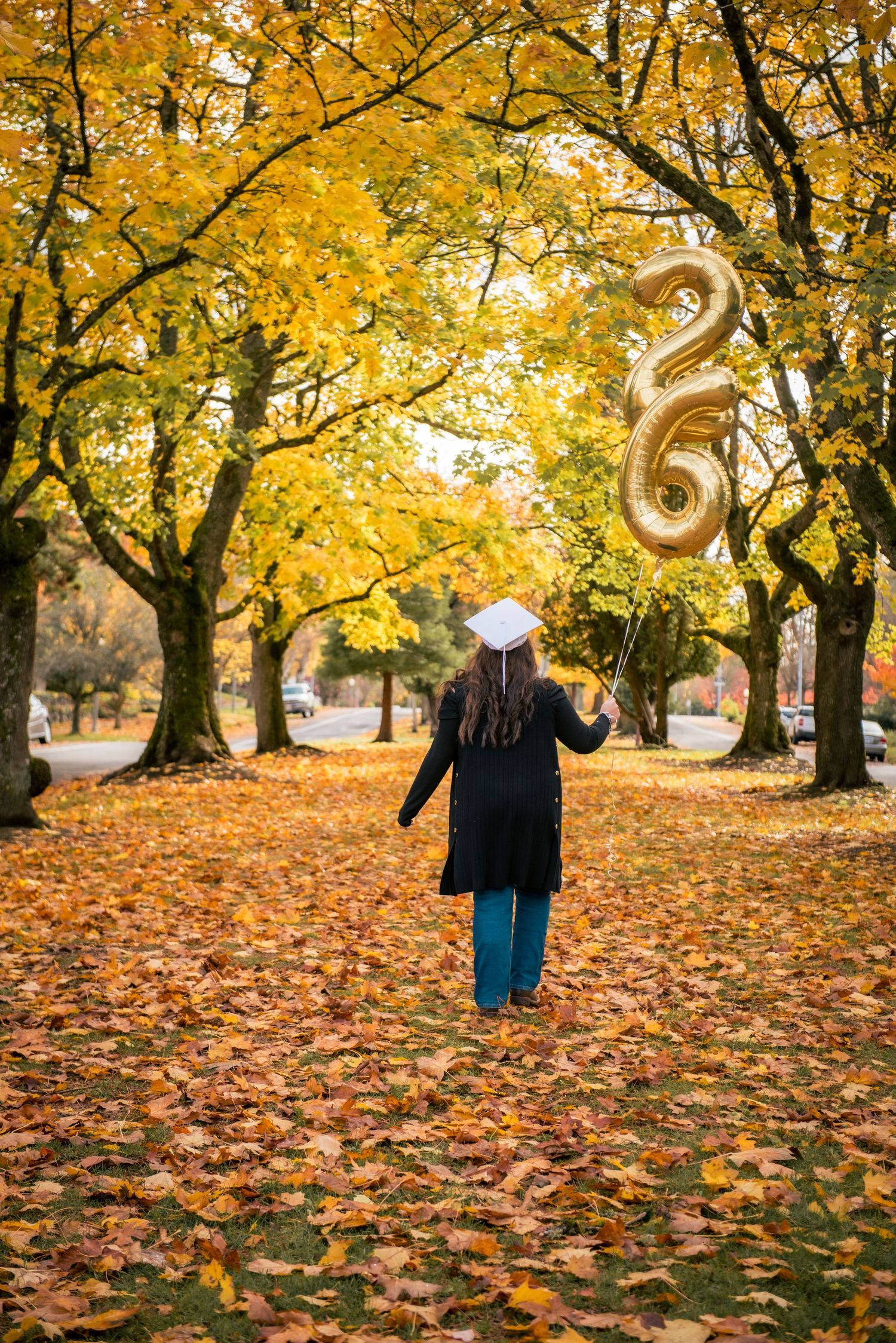 A figure holds a gold number 26 balloon while walking through a park filled with golden autumn leaves and trees.