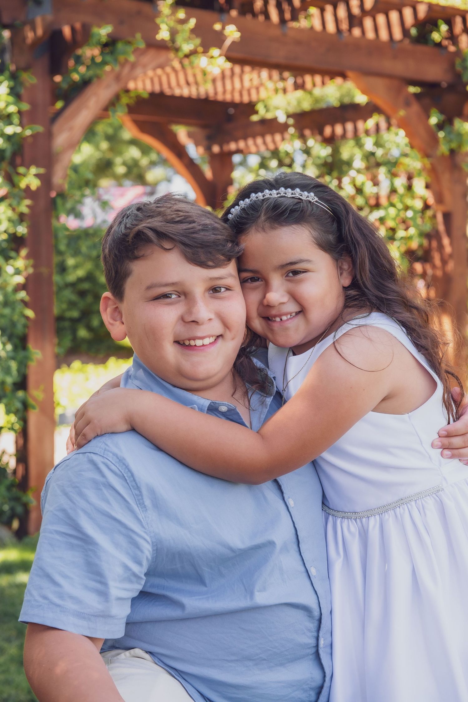 Two smiling children hugging outdoors under a wooden pergola, boy in blue shirt and girl in white dress with headband.