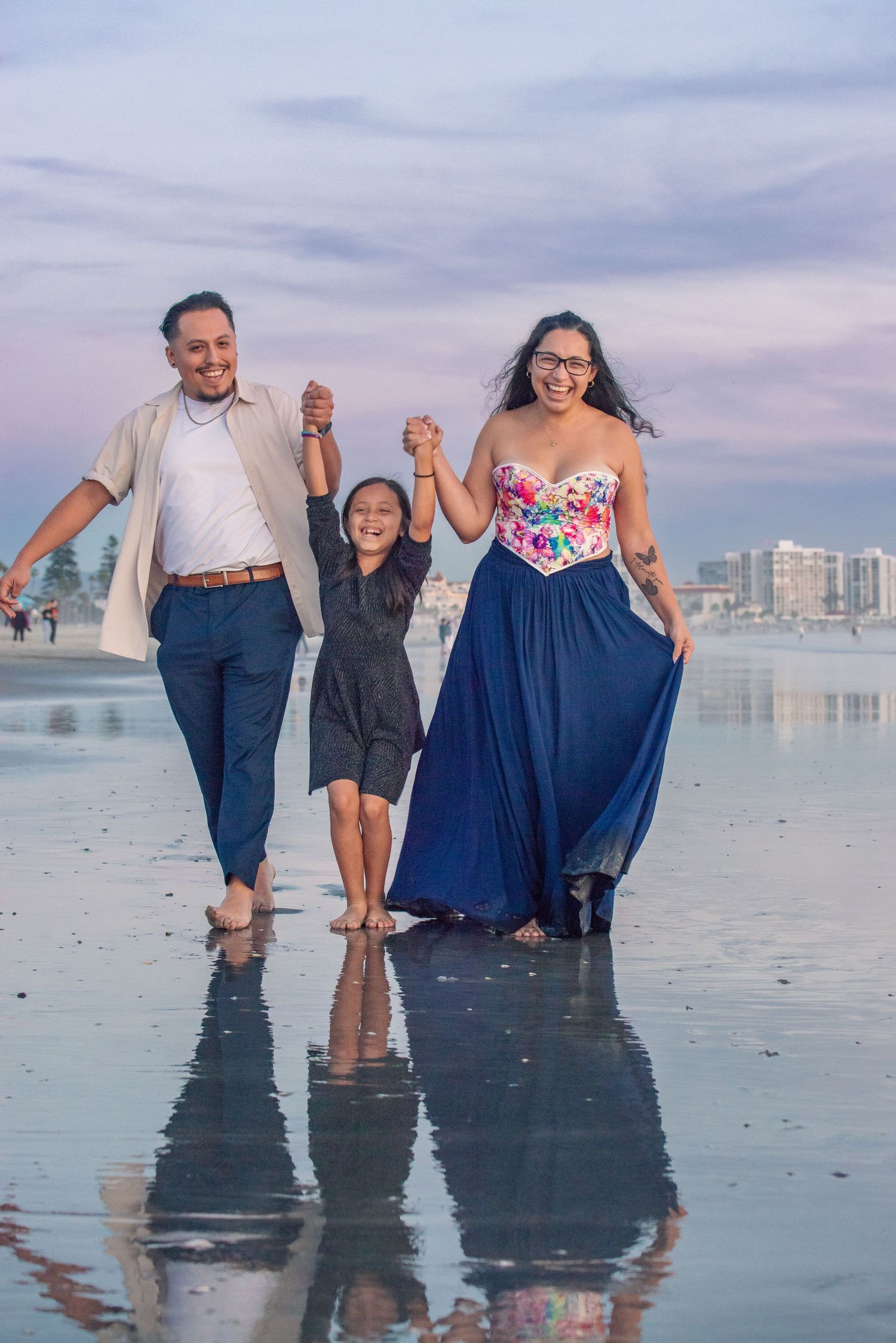 A family walks together holding hands on a reflective beach at sunset with city buildings in the background.