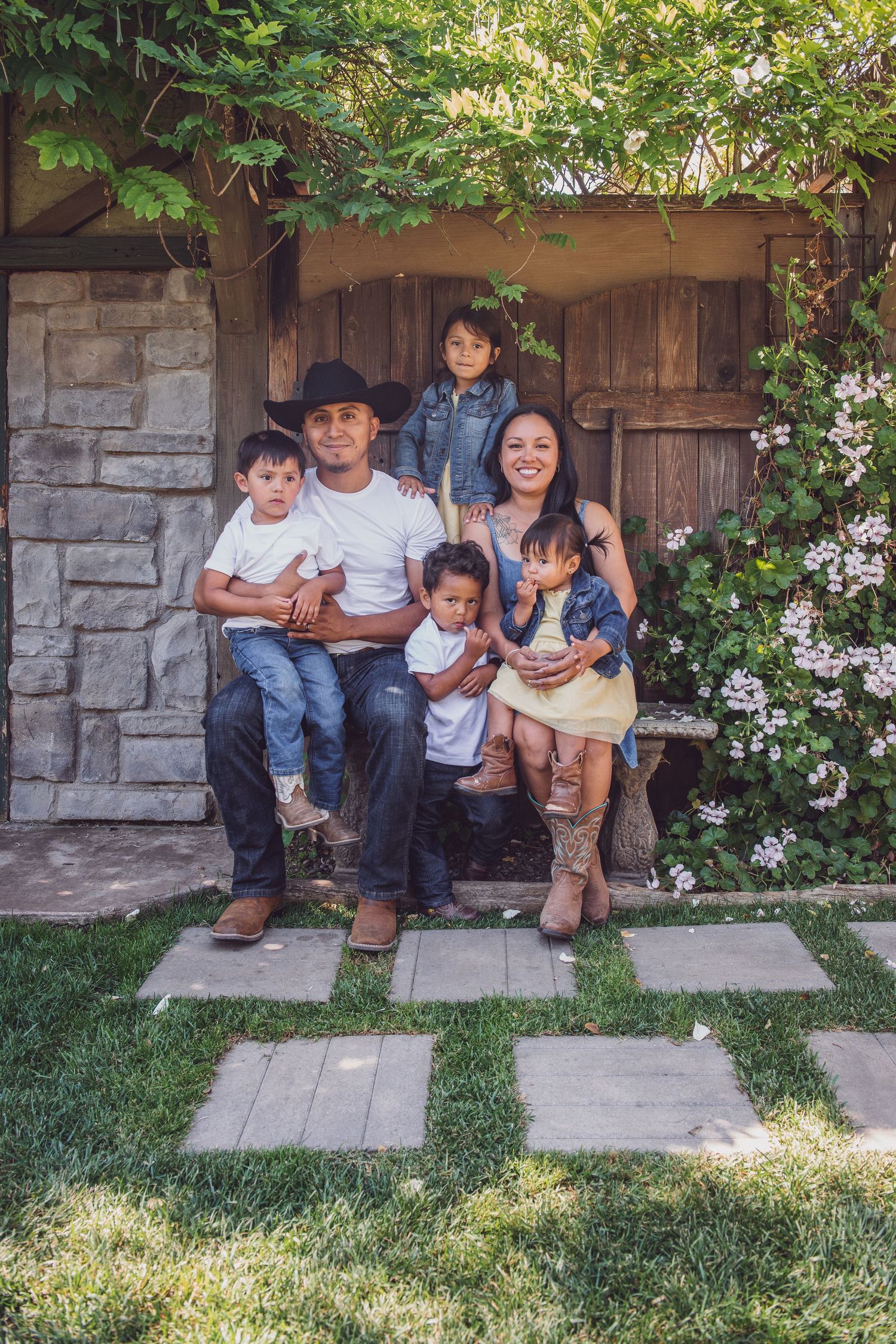 A family sits together on stone steps outside a rustic building surrounded by greenery and flowers.