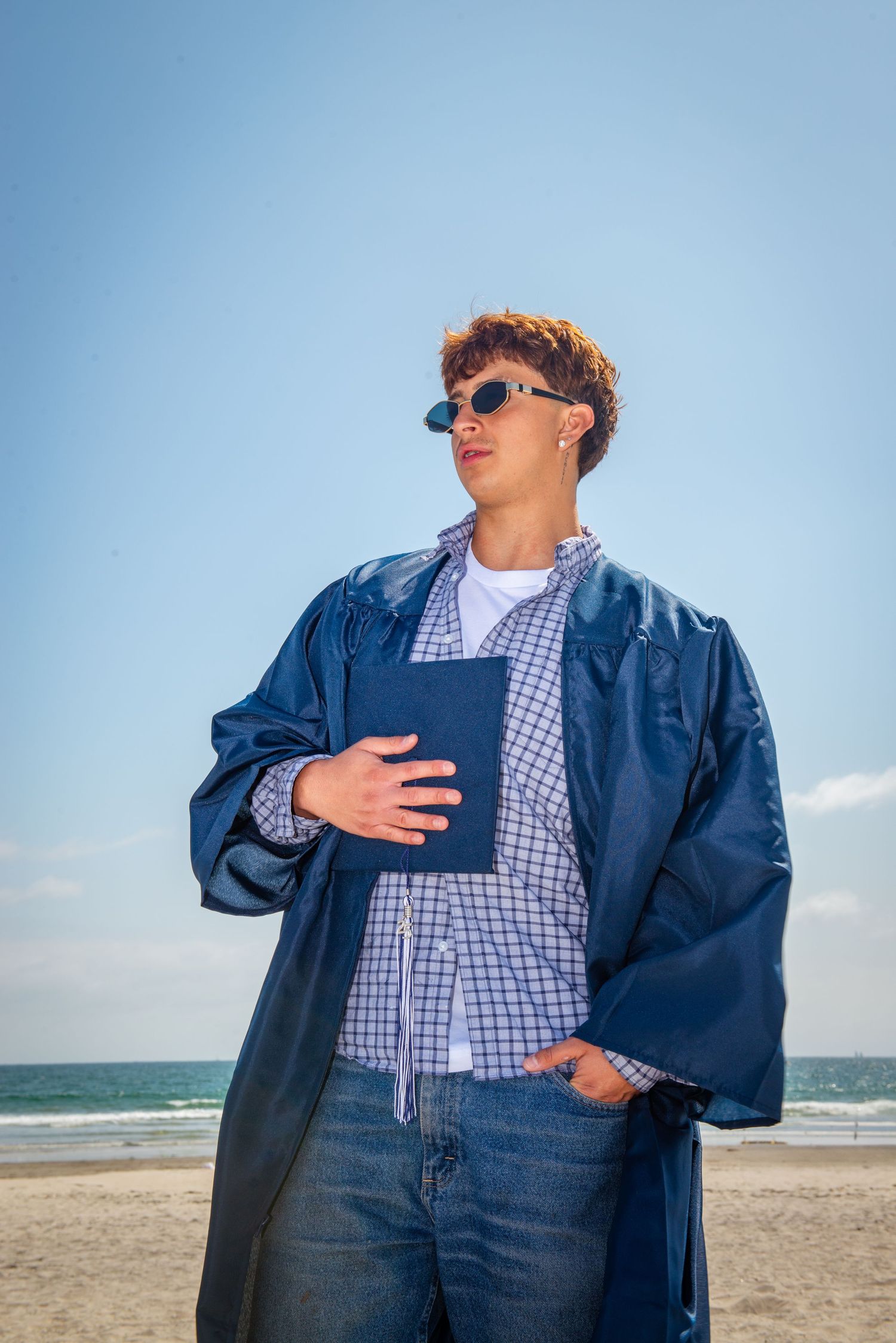 Person wearing navy blue jacket and gingham shirt poses against beach backdrop with ocean waves and blue sky.