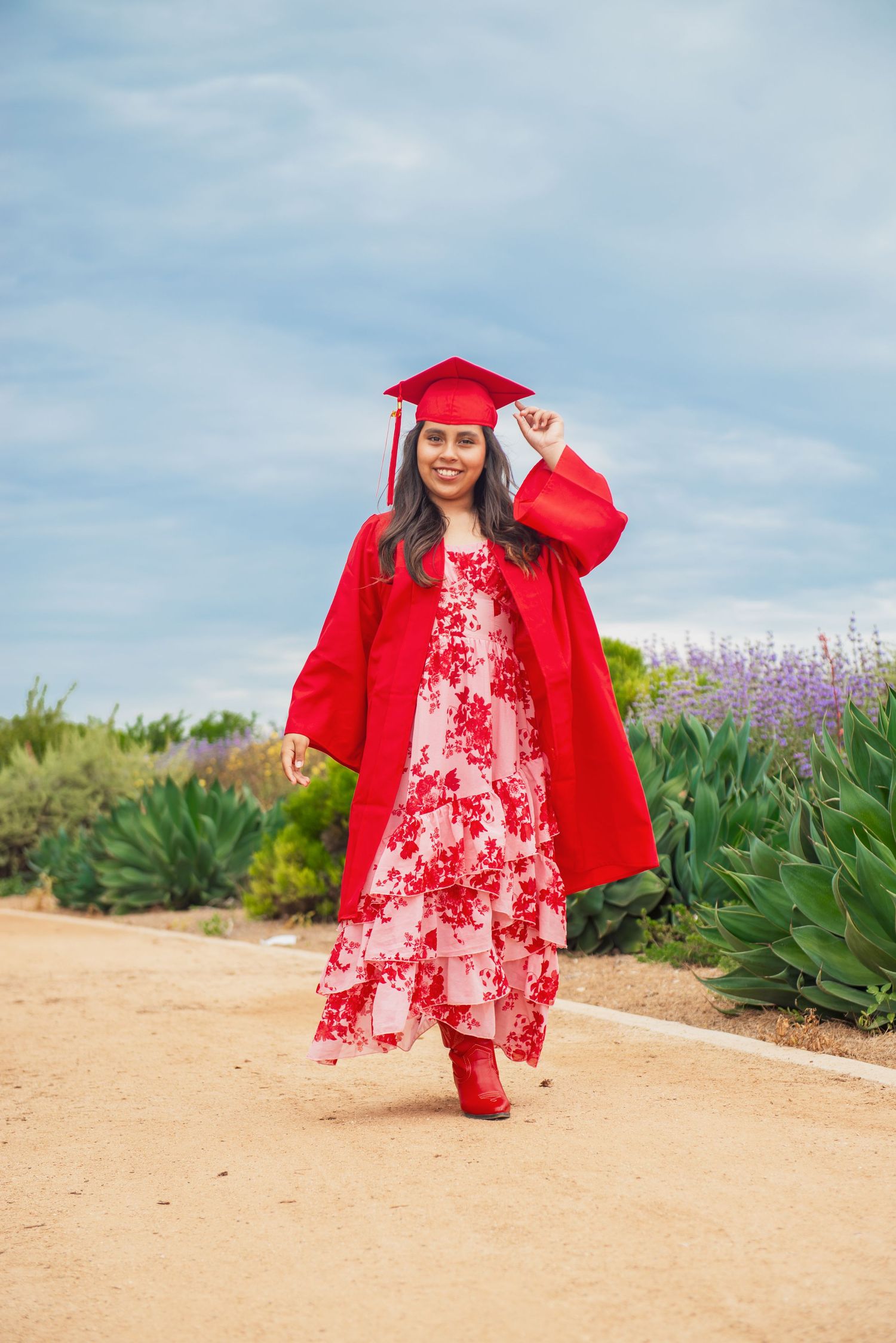 Graduate in red cap and gown poses on path with floral dress and matching heels against garden backdrop.