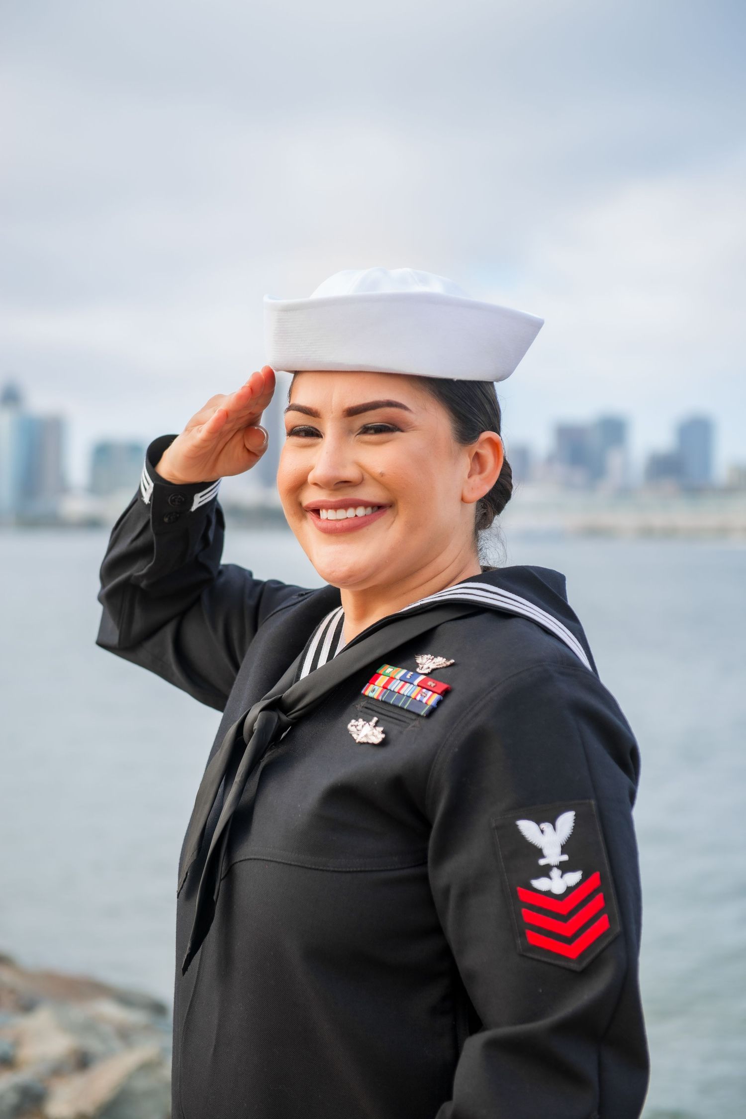 Navy service member in dress uniform salutes while standing near waterfront with city skyline in background.