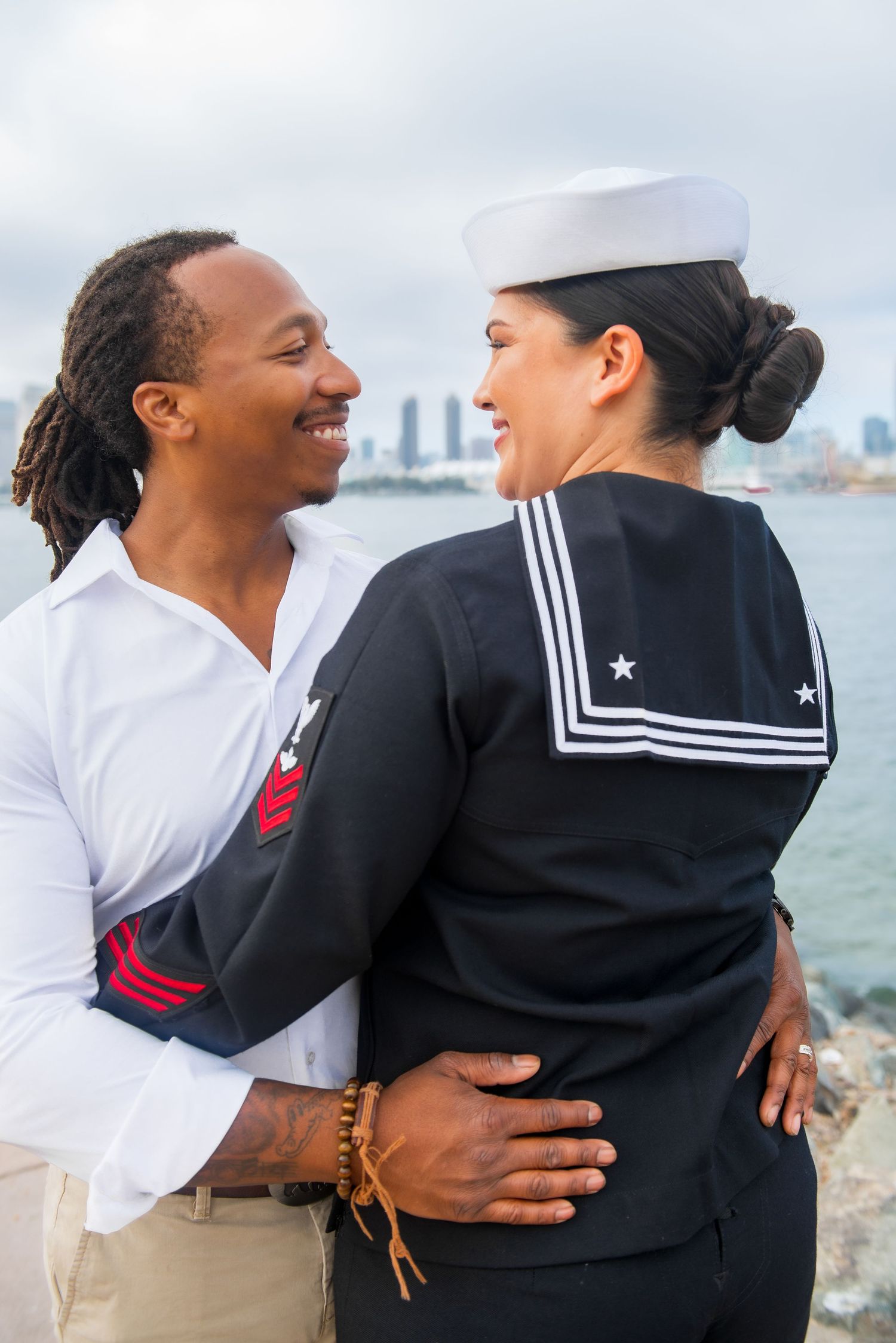 A couple shares an intimate moment by the waterfront as one wears a navy sailor uniform and the other wears a white shirt.