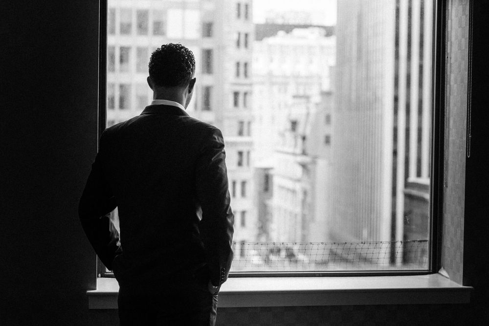 A silhouetted man handsomely dressed in his suit gazing out a window at city buildings.