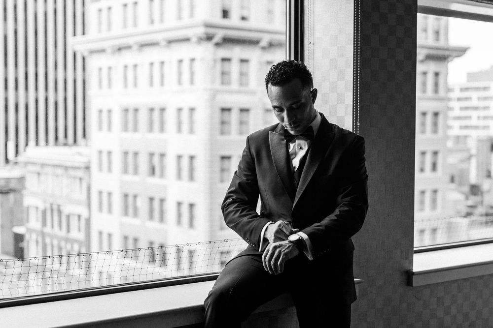 A black and white portrait of someone in formal attire by a window in an urban setting.