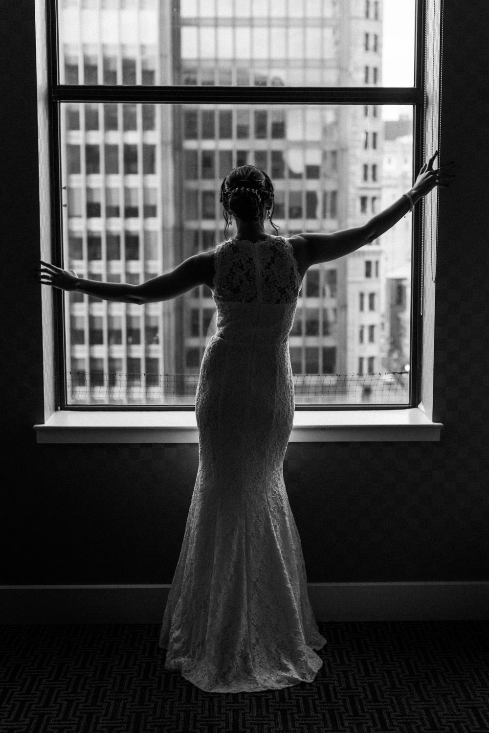 A dramatic silhouette of the bride in her wedding gown against a city window view.
