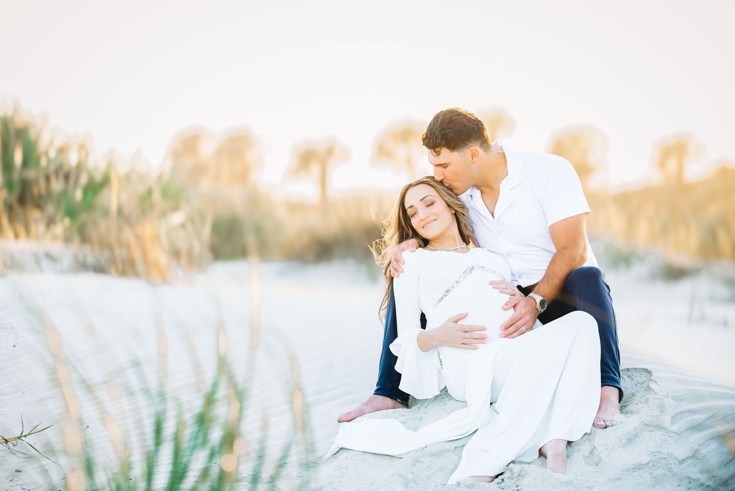 Couple sitting on beach sand dunes, woman in white dress with partner embracing her during maternity photo session.