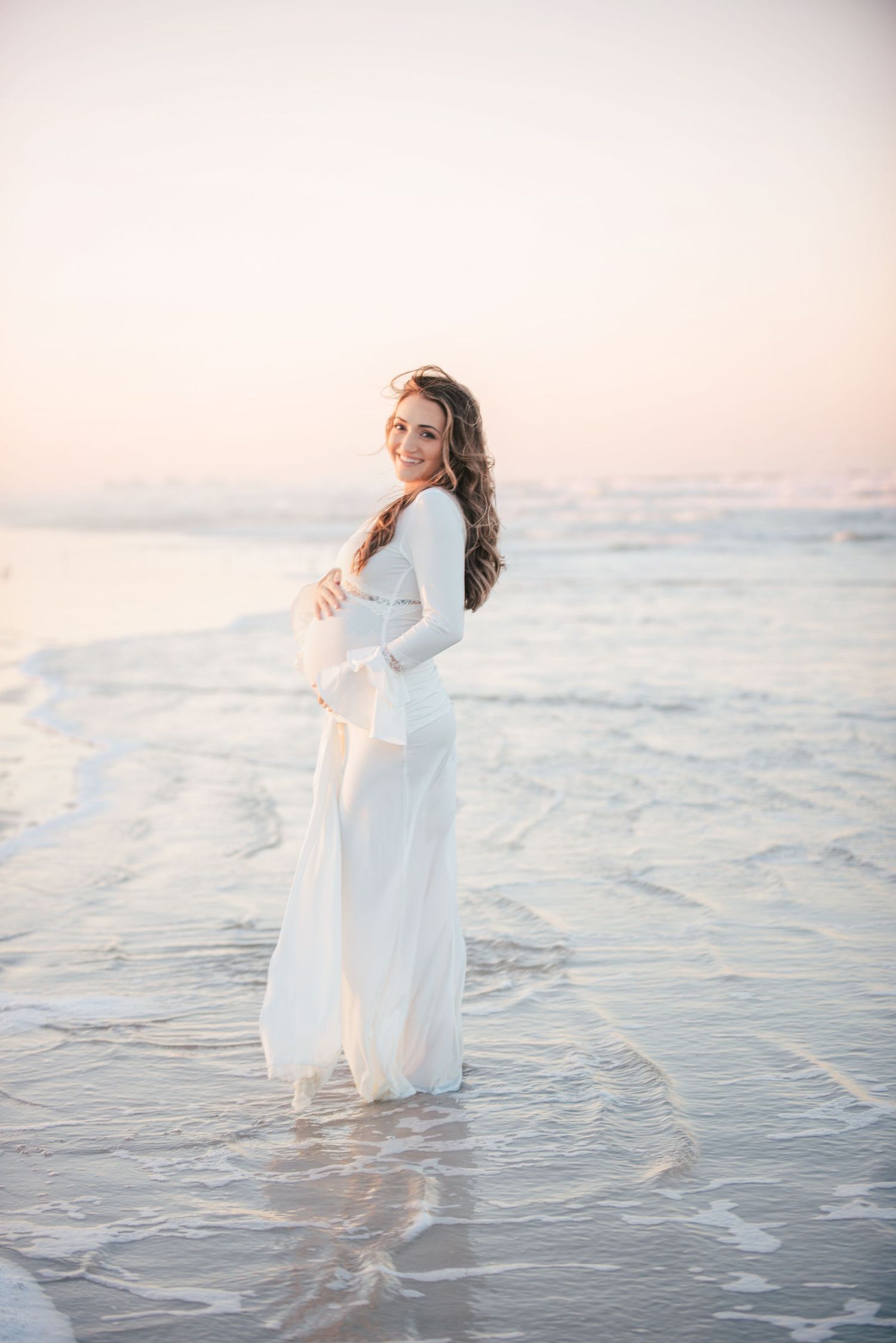 Pregnant woman in white dress standing in ocean waves at sunset beach, smiling over shoulder.