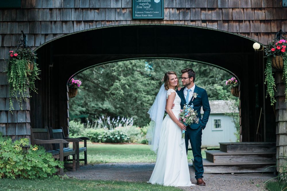 Couple in wedding attire embracing under a rustic wooden structure surrounded by greenery.