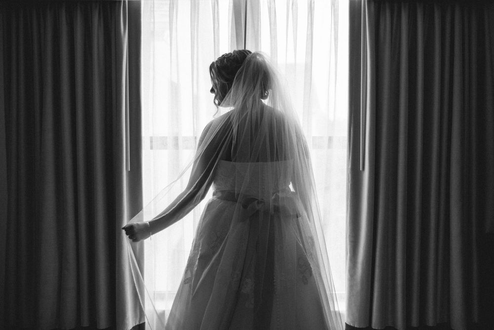 Silhouette of a bride's dress against white curtains in dramatic lighting.