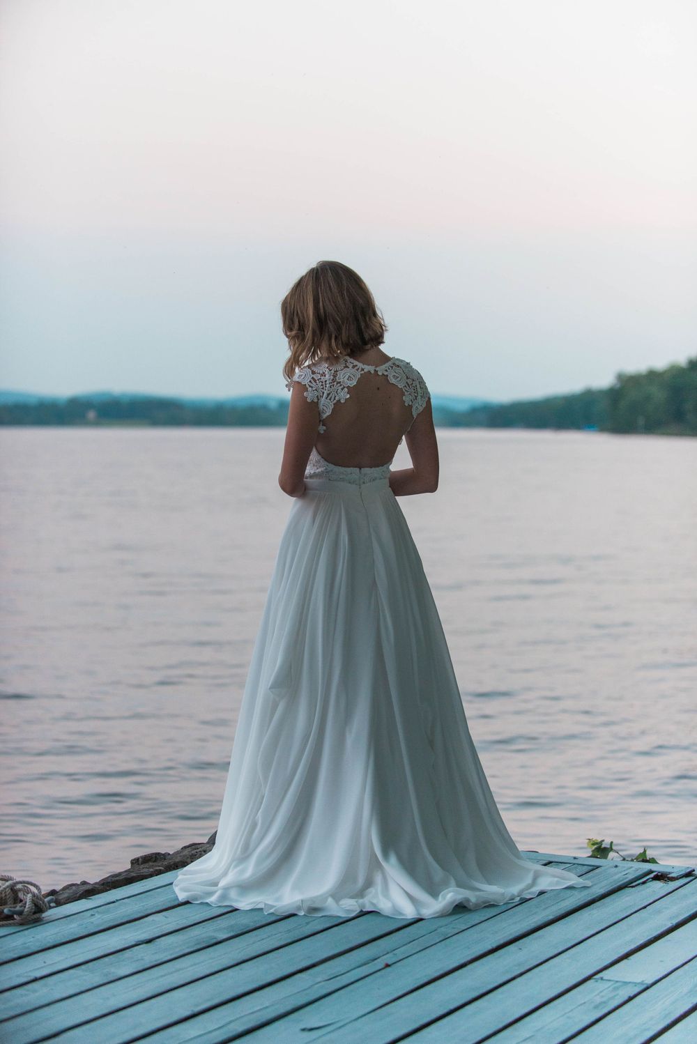 A bride in her flowing white dress stands on a dock overlooking a serene lake at dusk.