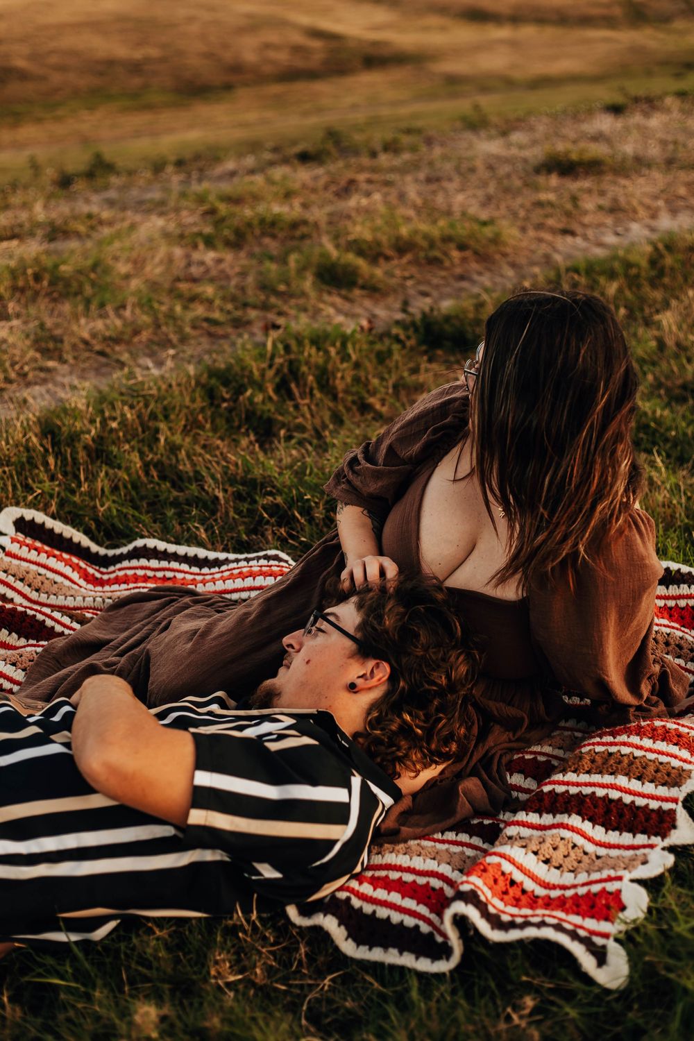 Two people lying together on a striped blanket outdoors in a field during sunset.