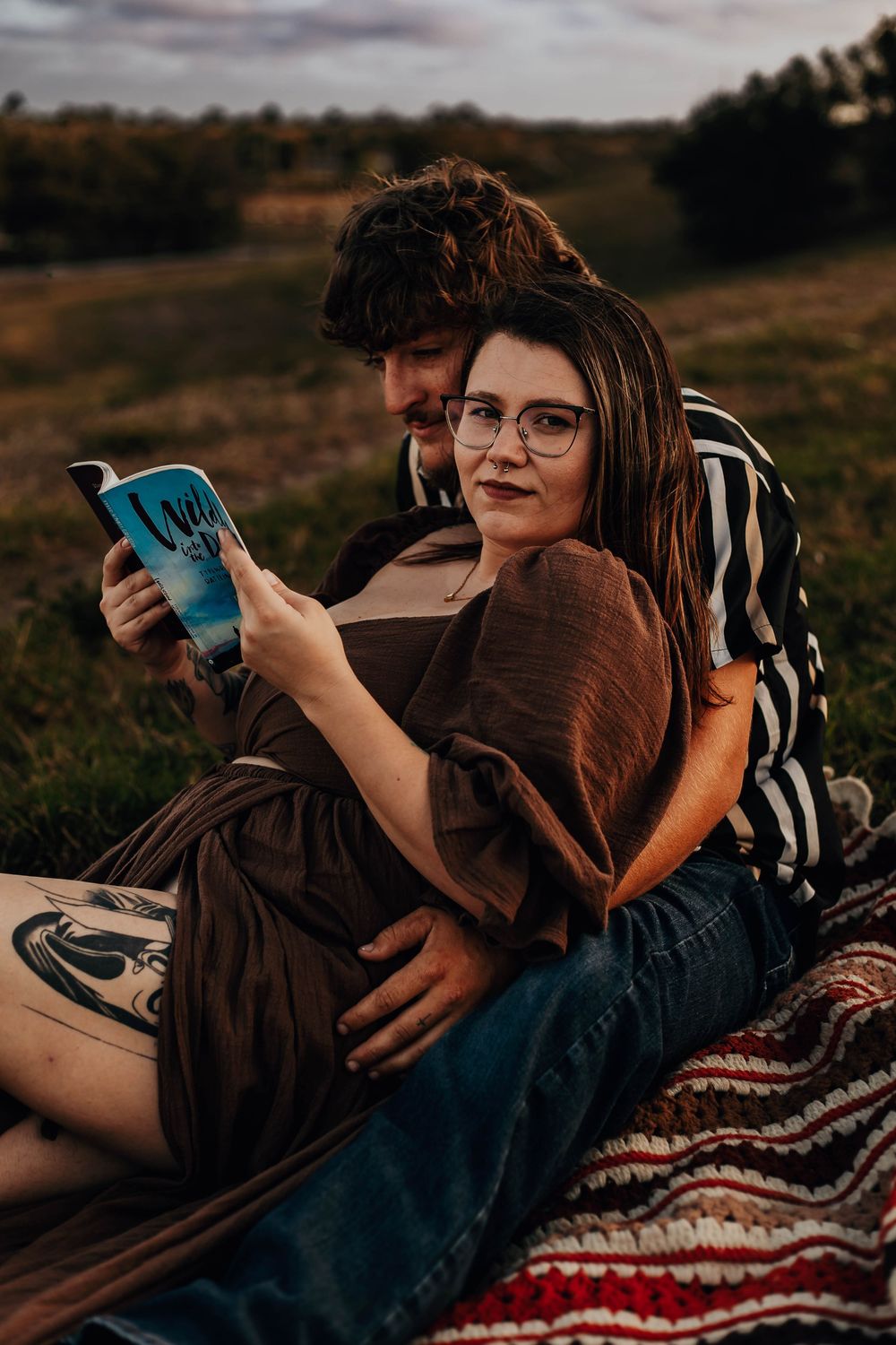 A couple relaxes on a blanket with a striped pattern during golden hour in a rustic outdoor setting.